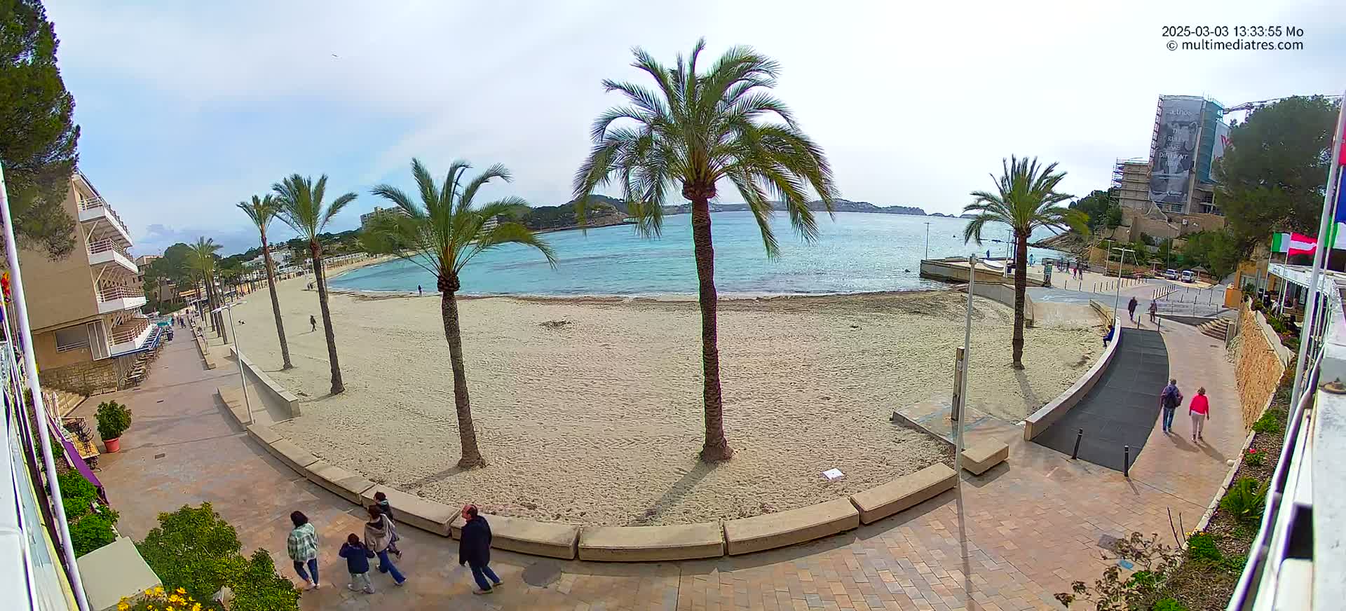 A mostly empty sandy beach with palm trees and a paved walkway is seen on a partly cloudy day, overlooking calm blue water.