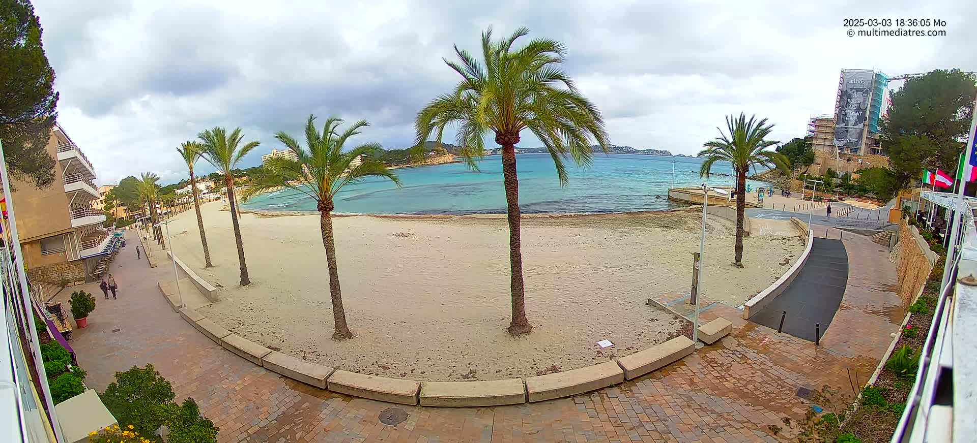 A sandy beach with palm trees lines a paved walkway leading to a calm, turquoise ocean under a cloudy sky.