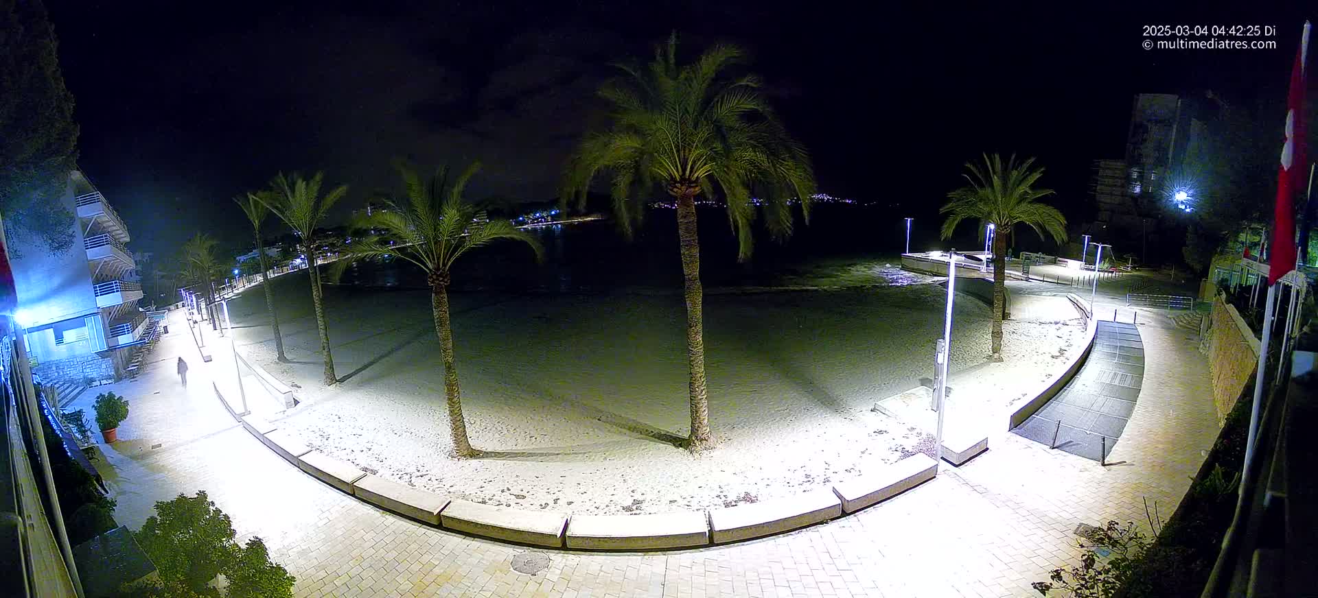 A nighttime, brightly lit beach scene shows a sandy beach, palm trees, and a paved walkway curving along the shore, with buildings visible in the background.