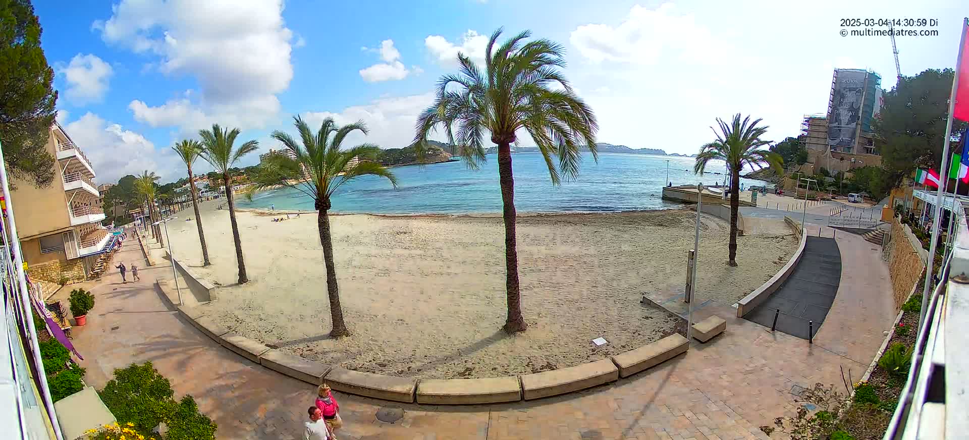 A sandy beach with palm trees and a paved walkway, overlooking calm blue water under a partly cloudy sky.