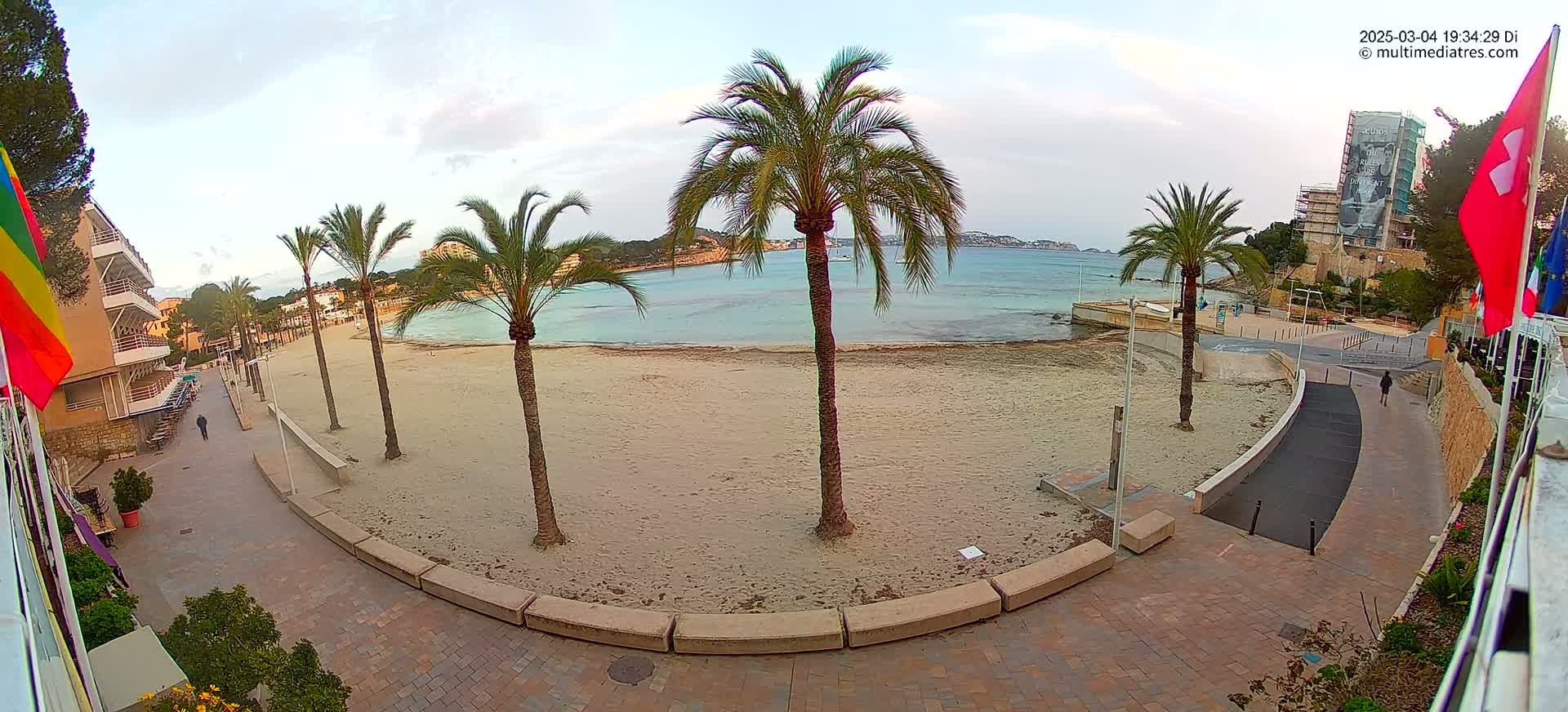 A mostly empty sandy beach with palm trees is seen from an elevated viewpoint, under a mostly cloudy sky.