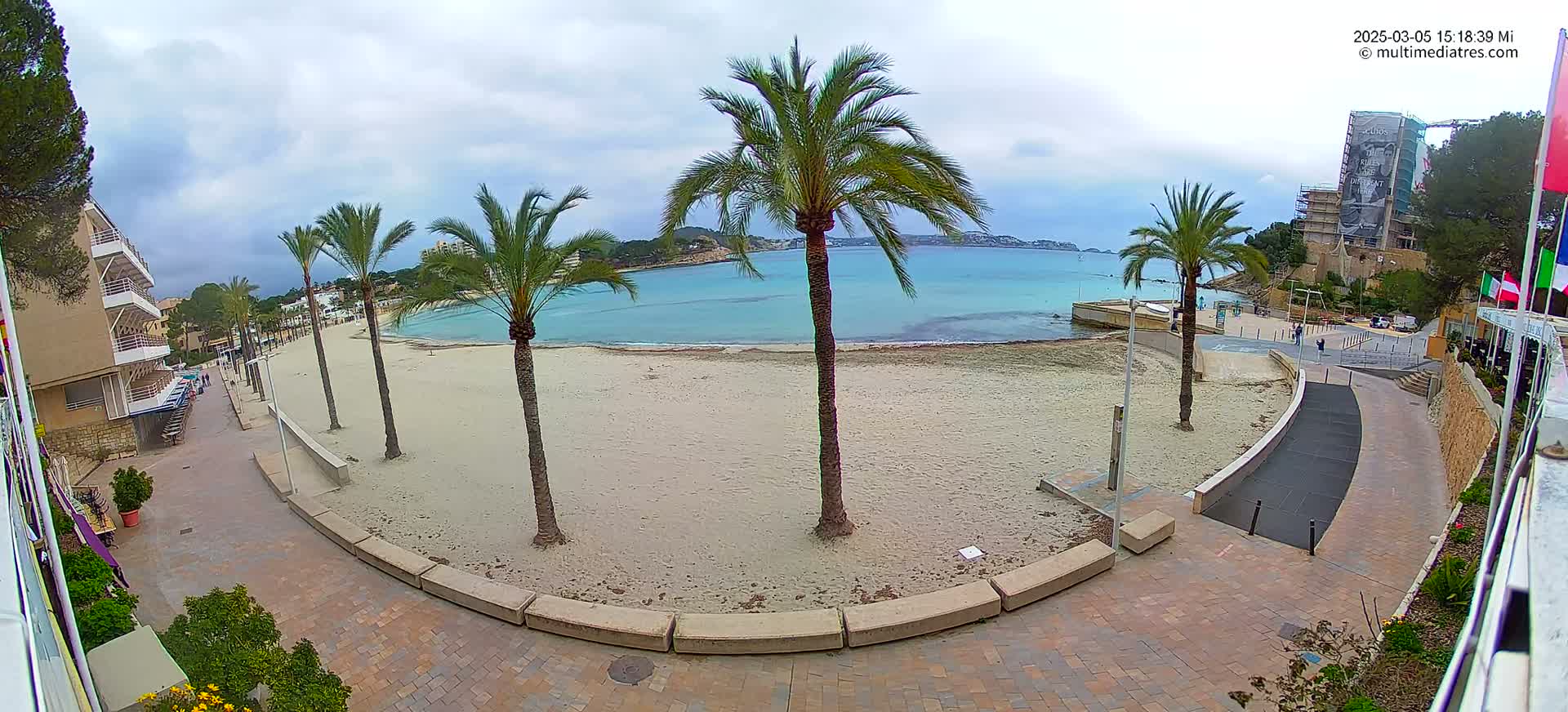 A sandy beach with palm trees lining a paved walkway next to calm, turquoise water under an overcast sky.