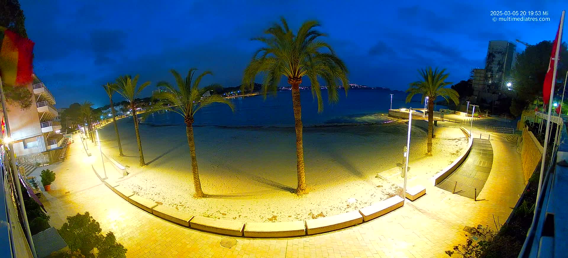 An illuminated sandy beach at night, bordered by palm trees and a paved walkway, overlooks a calm body of water under a dark blue twilight sky.