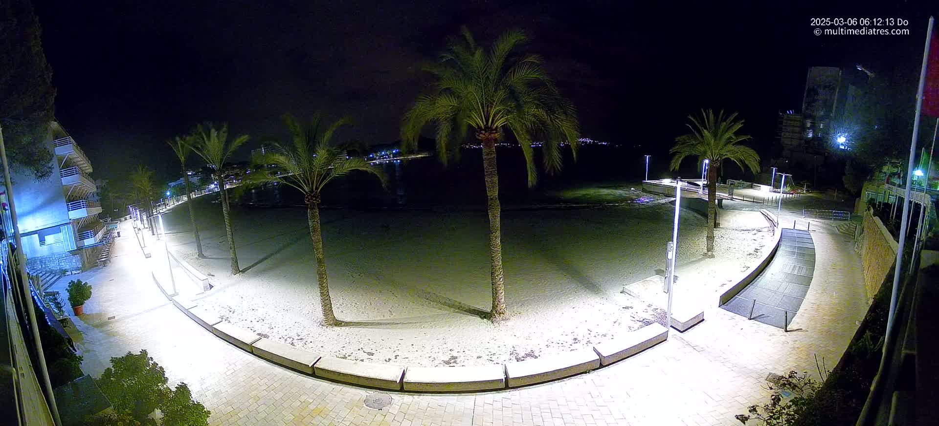A nighttime, illuminated view of a sandy beach with palm trees, a paved walkway, and buildings in the background.