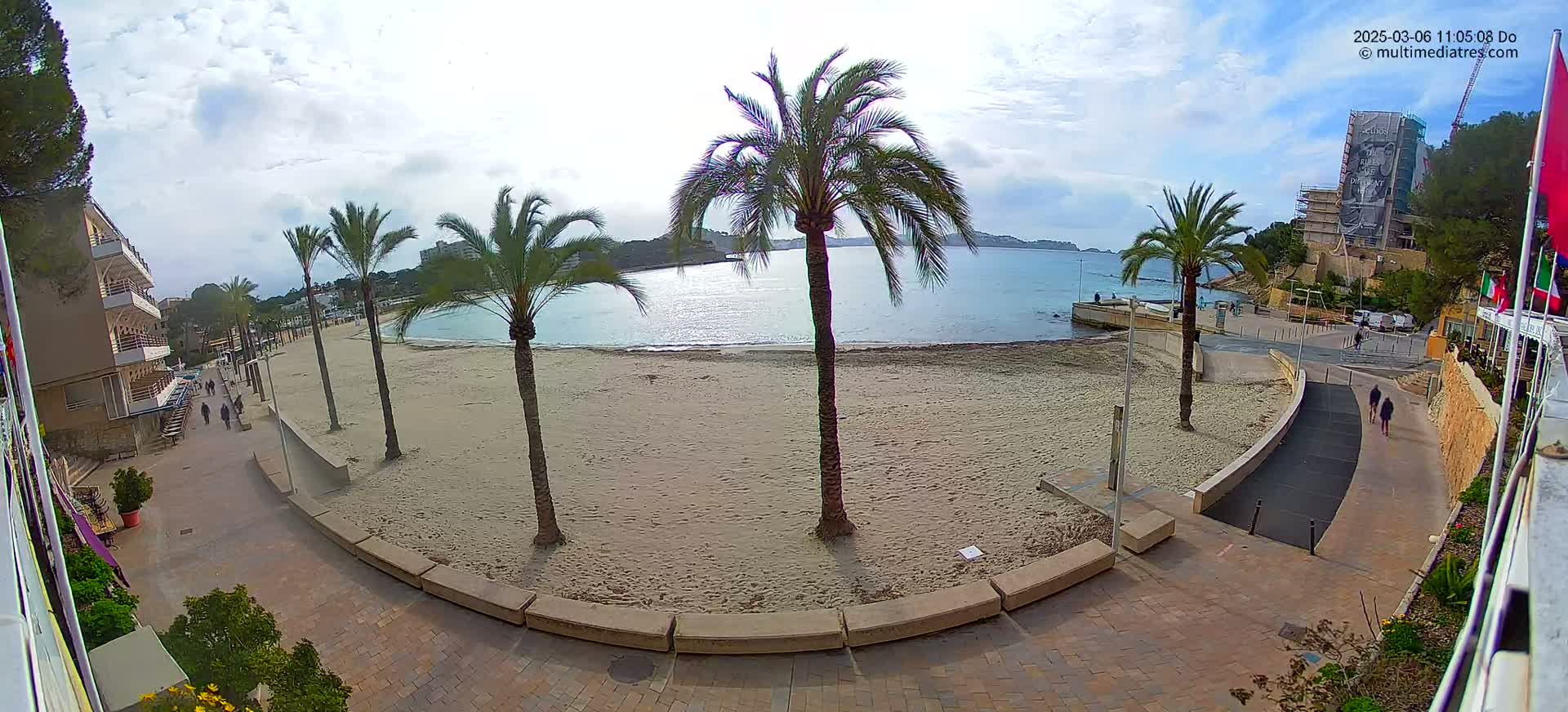 A sandy beach with palm trees, a calm sea, and a paved walkway leading to a building, under a partly cloudy sky.