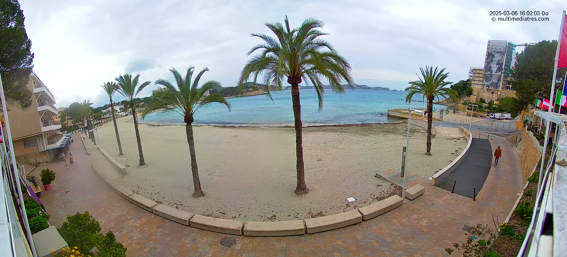 A sandy beach with several palm trees is visible, next to a paved walkway leading to a calm, light-blue sea under an overcast sky.