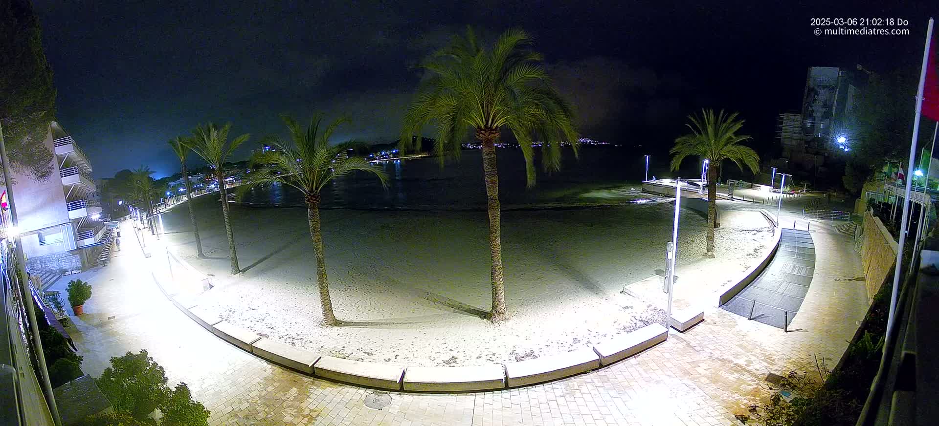 A nighttime view of a sandy beach at low tide, lined with palm trees and illuminated walkways, under a clear, dark sky.