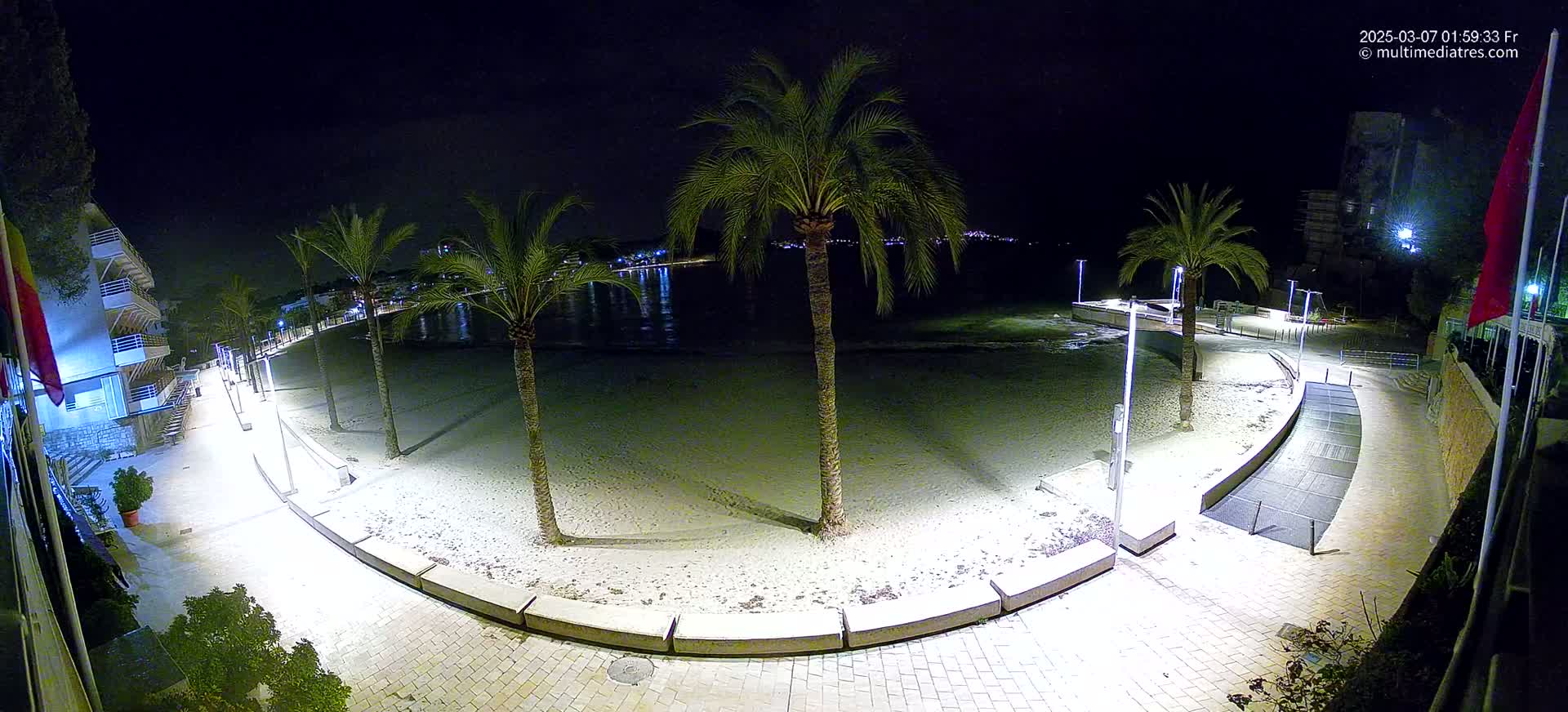 A nighttime, illuminated view of a sandy beach bordered by a paved walkway lined with palm trees and buildings.