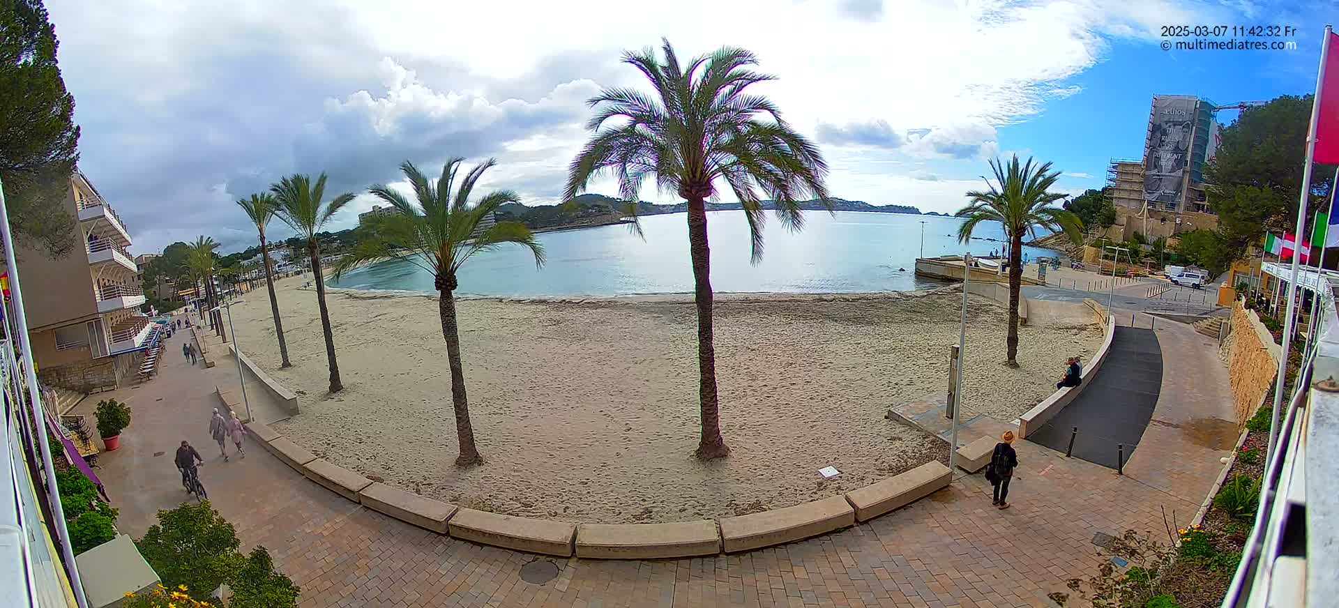 A mostly sunny day shows a sandy beach with palm trees and a few people walking along a paved walkway next to a calm body of water.