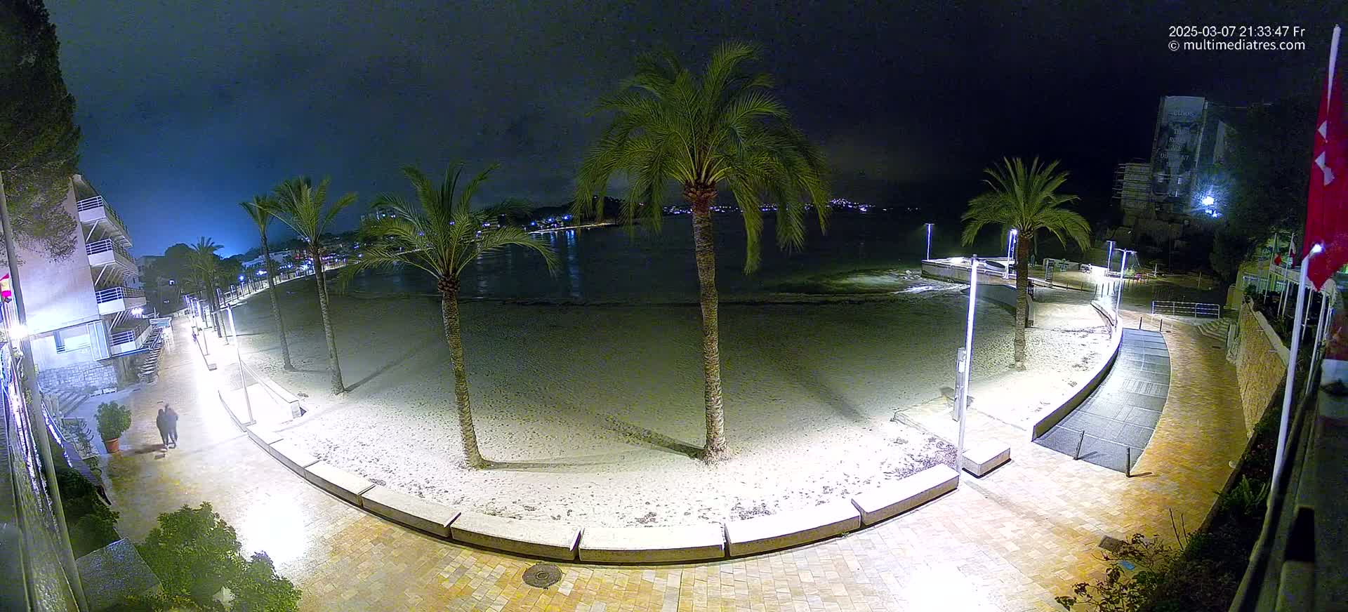A nighttime view of a mostly empty sandy beach, bordered by a paved walkway lined with palm trees and buildings, under a dark, clear sky.