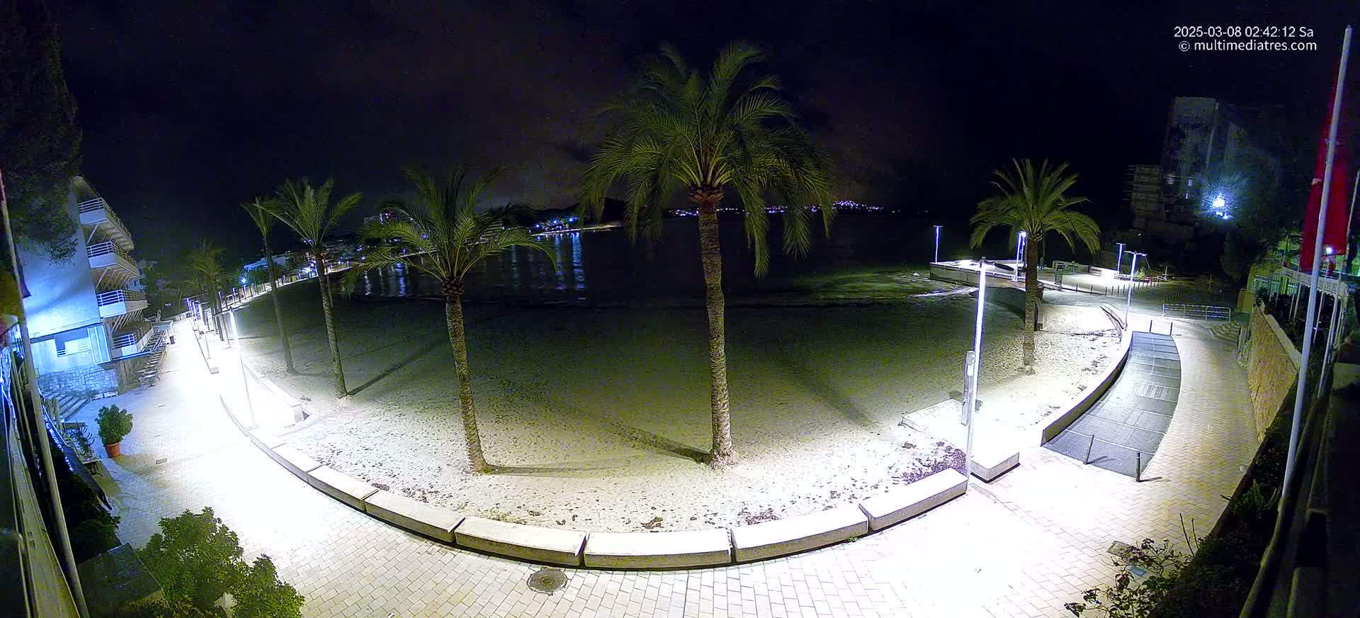 A nighttime, illuminated view of a sandy beach bordered by a paved walkway, palm trees, and buildings, overlooking a calm body of water.