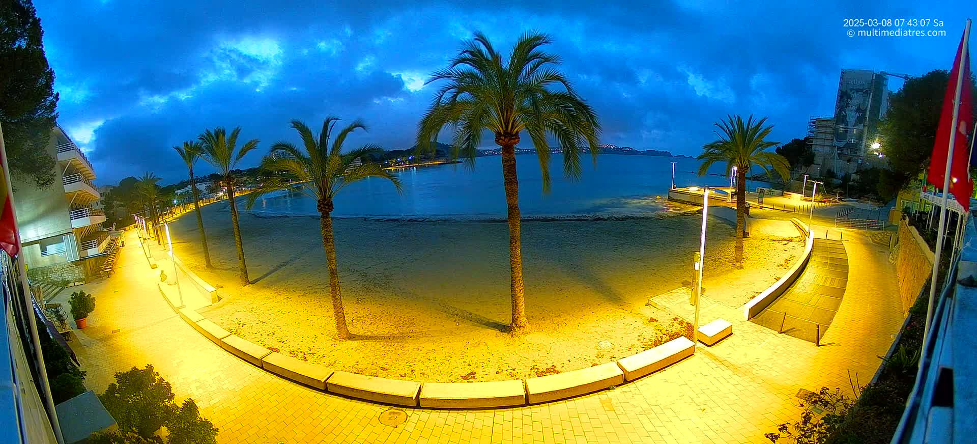 A nighttime beach scene under a partly cloudy blue sky shows a sandy beach with palm trees, illuminated walkways, and buildings in the background.