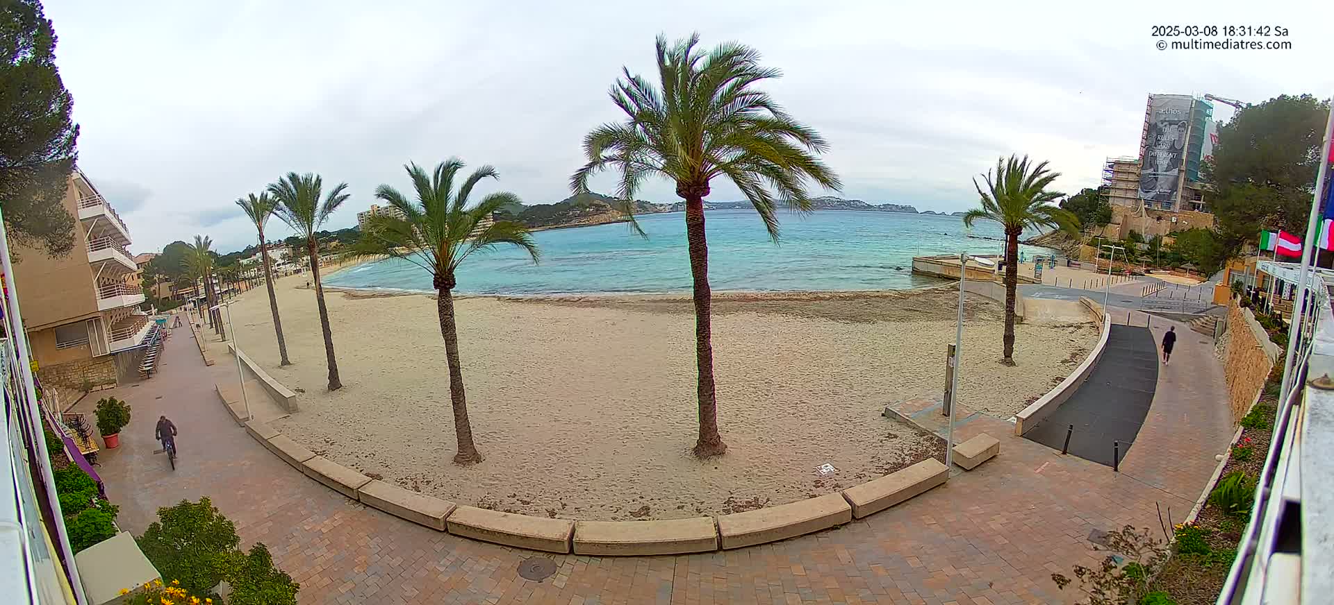 A sandy beach with palm trees and a paved walkway, overlooking calm turquoise water under an overcast sky.