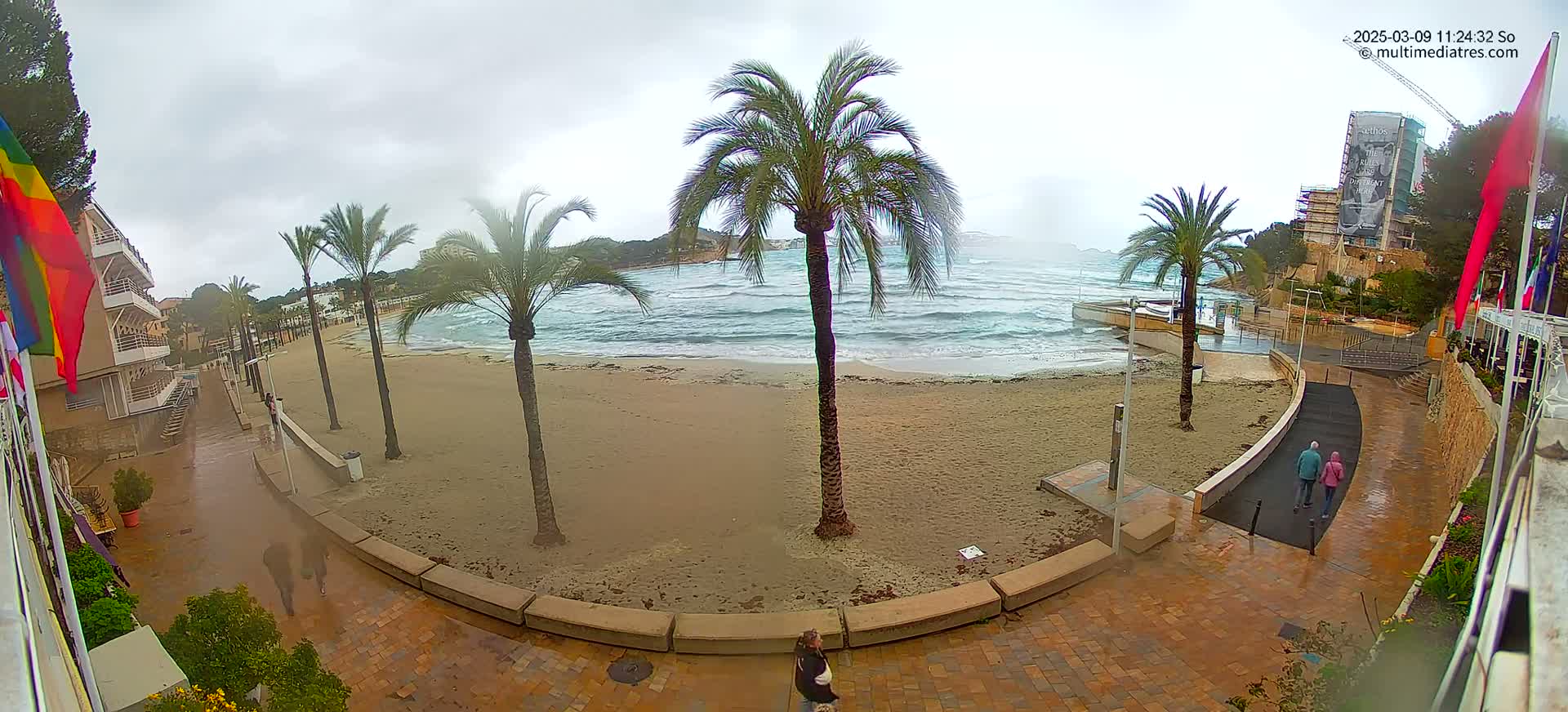 A sandy beach with palm trees is seen from an elevated viewpoint on an overcast day, with choppy waves in the ocean and people walking along a paved pathway.