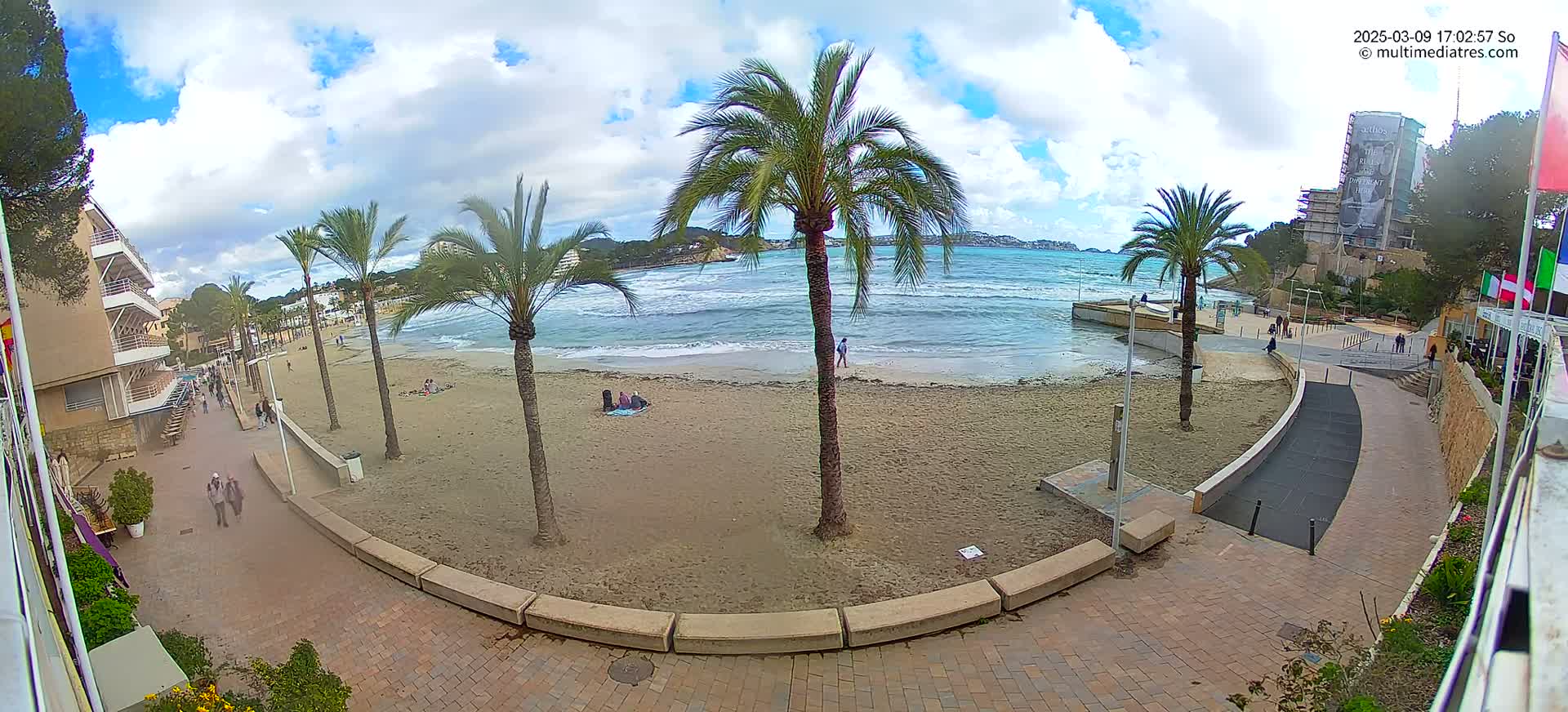 A sandy beach with palm trees, a few people, and some buildings is visible under a partly cloudy sky.