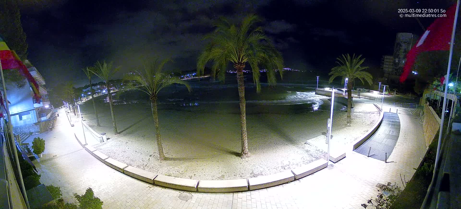 A nighttime view of a sandy beach with palm trees, gently lapping waves, and illuminated walkways.