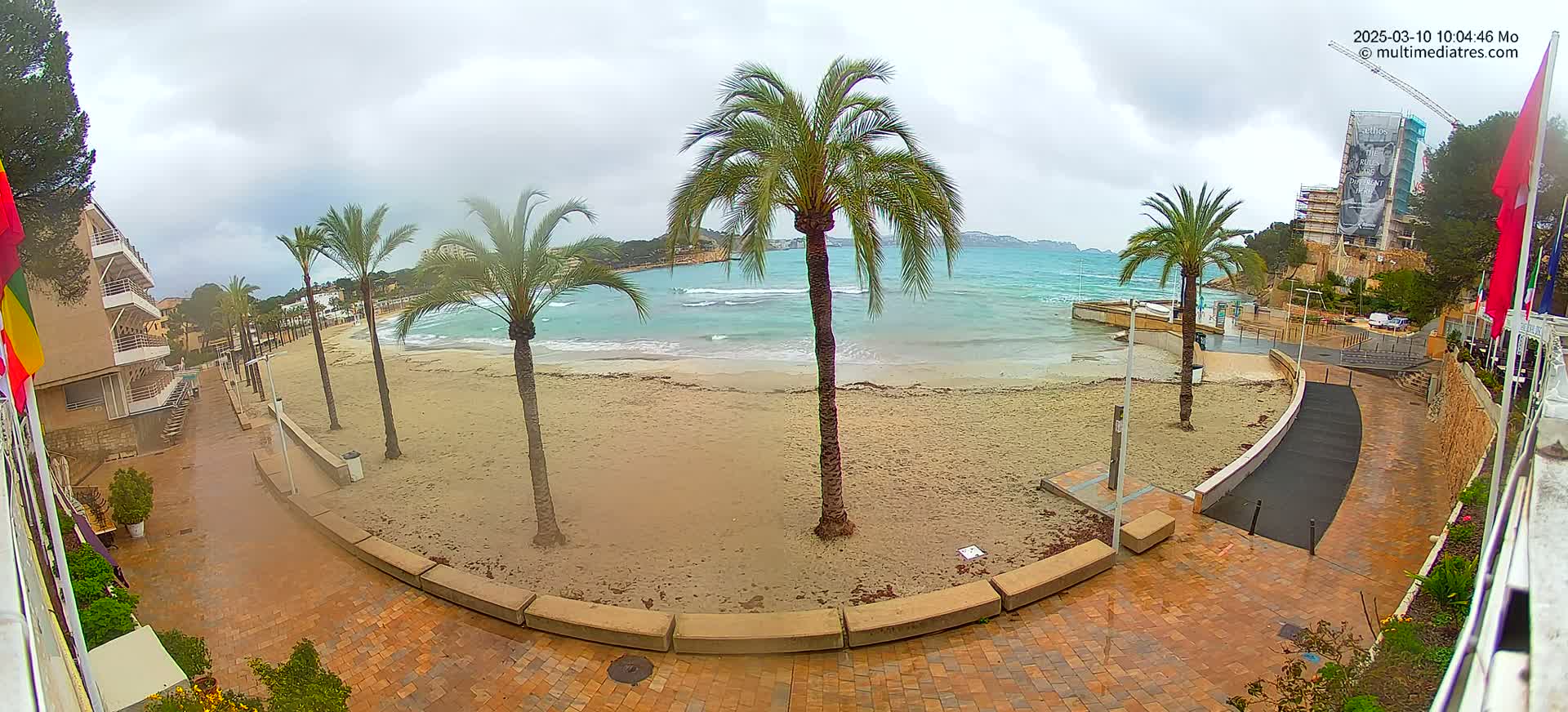 A sandy beach with palm trees is visible from a waterfront promenade on an overcast day.
