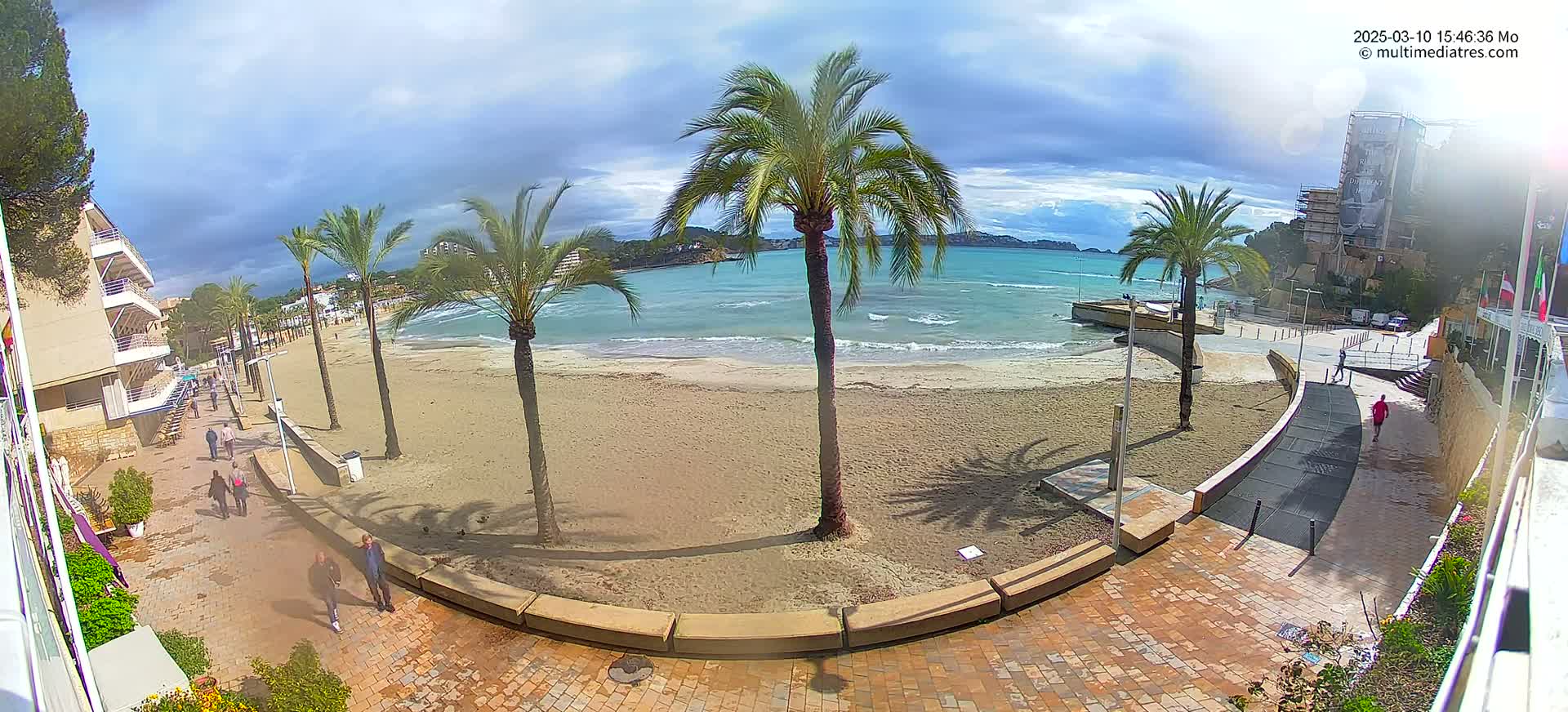 A sandy beach with palm trees and several people walking along a paved walkway, overlooking a calm, somewhat wavy ocean under a partly cloudy sky.