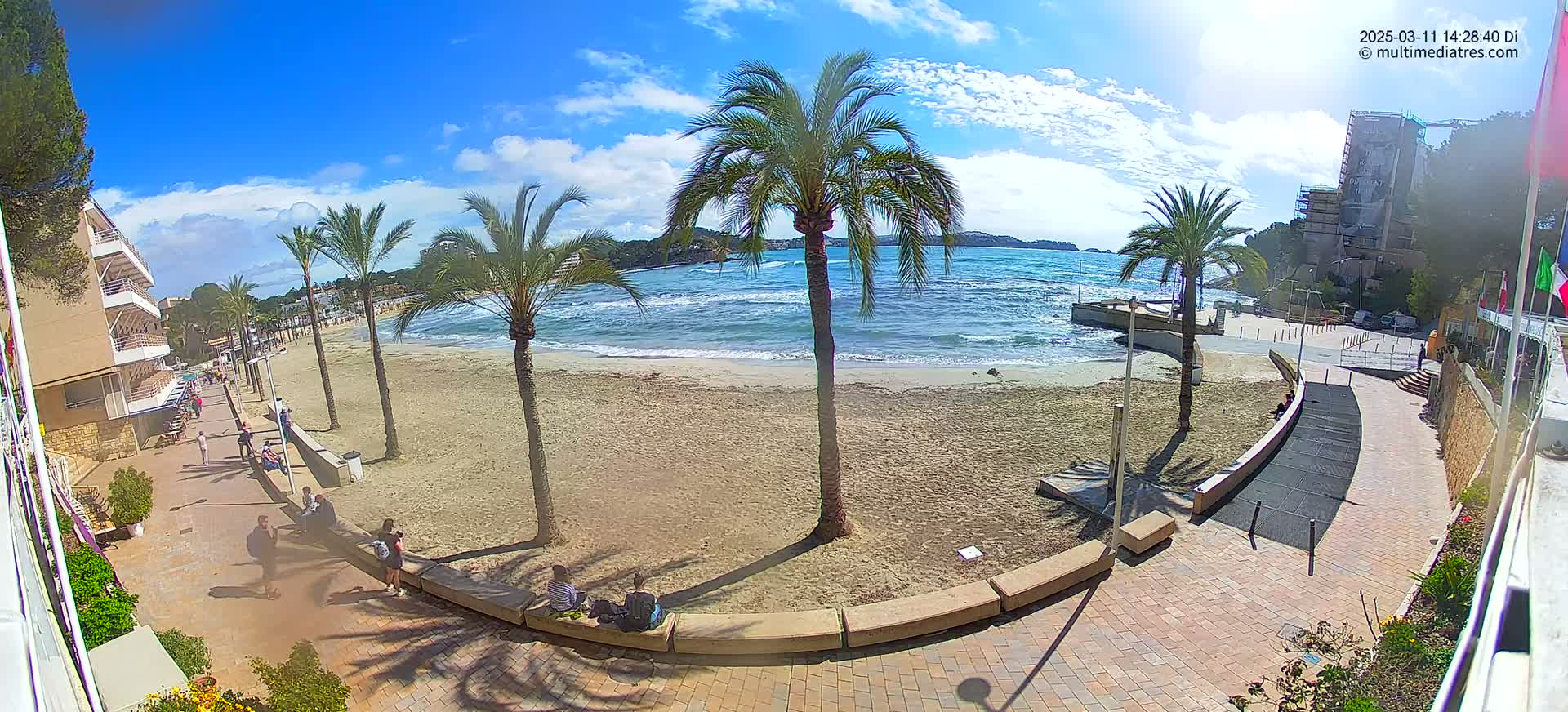 A sandy beach with palm trees and a few people is seen under a mostly sunny sky with some clouds, next to a paved walkway and buildings.