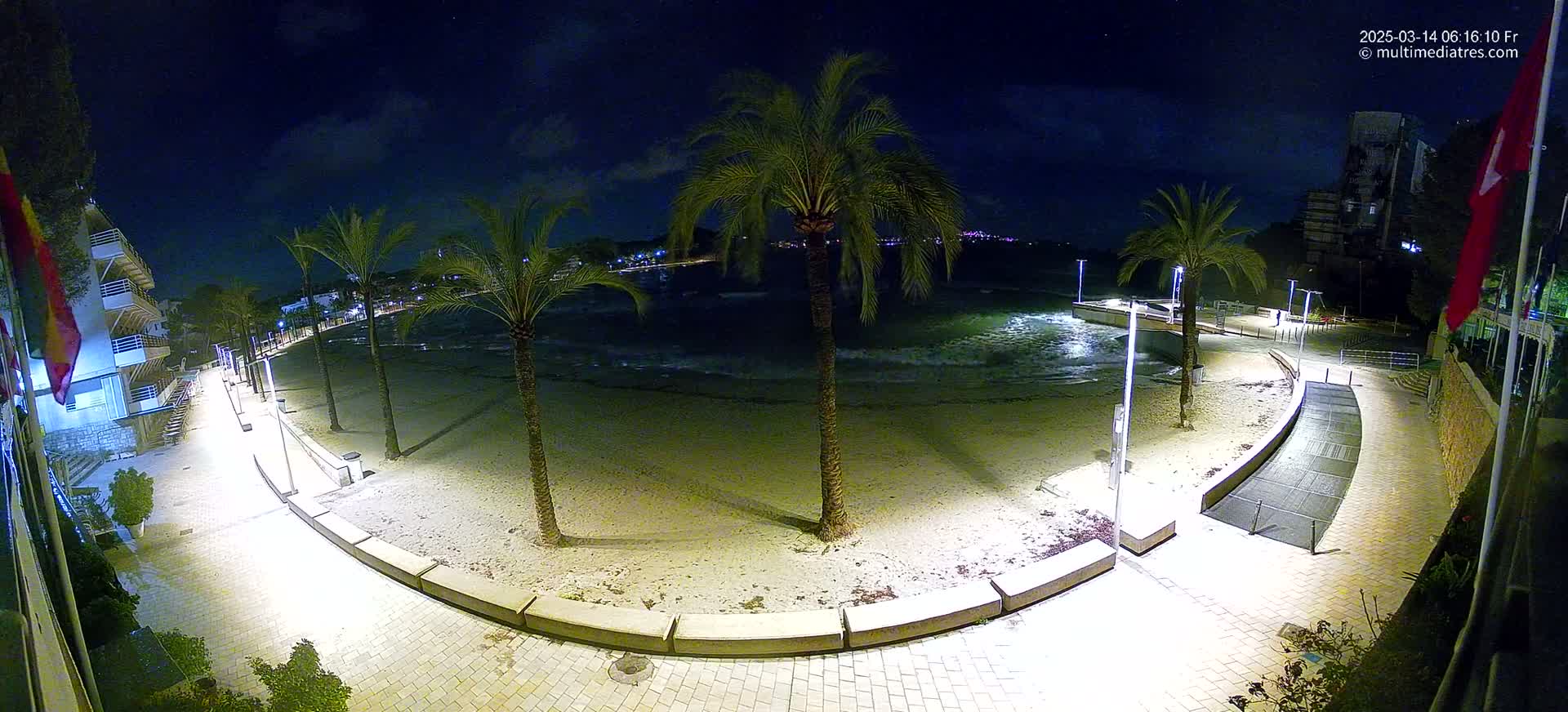 A nighttime, illuminated view of a sandy beach with palm trees, a paved walkway, and gentle waves lapping the shore.