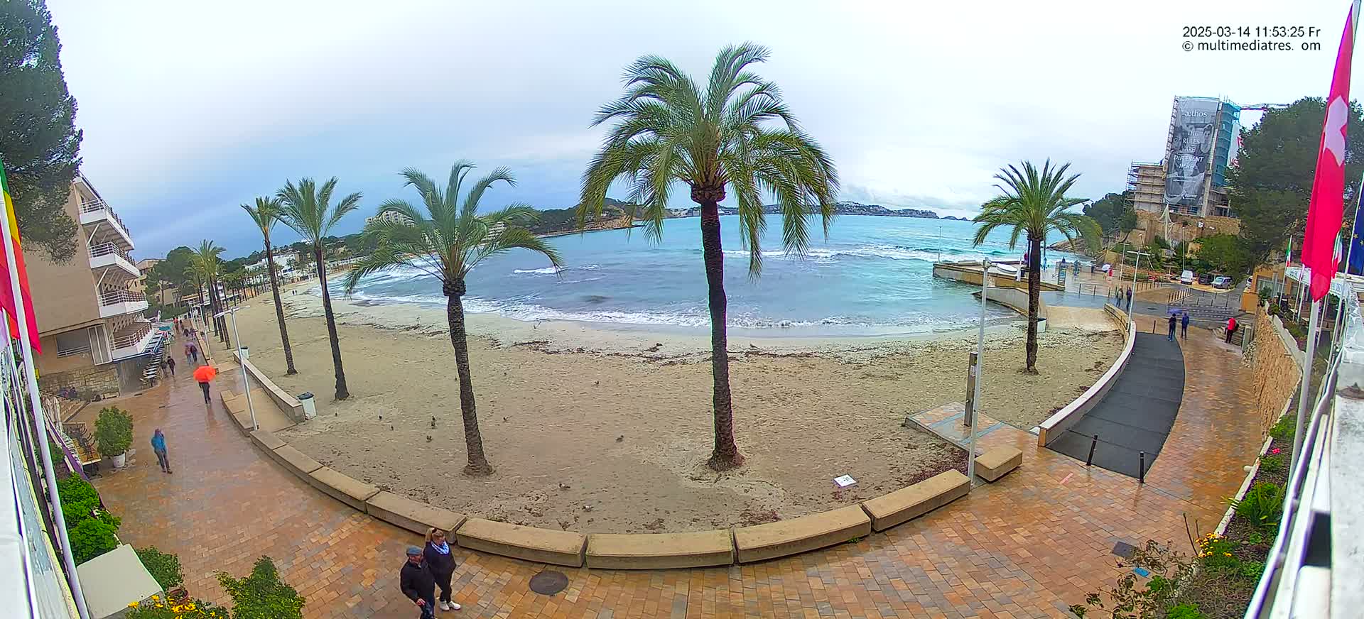 A sandy beach with palm trees and several people walking along a paved walkway, overlooking a somewhat choppy sea under an overcast sky.