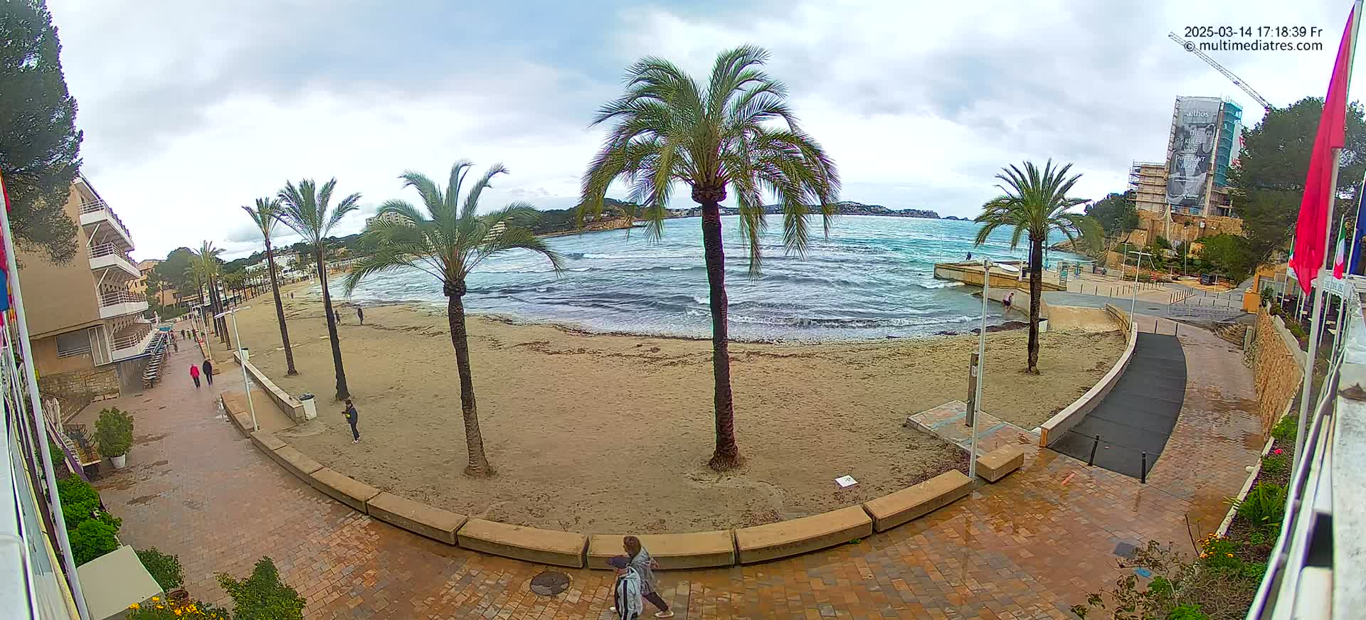 A sandy beach with palm trees and buildings is visible under an overcast sky, with a somewhat choppy sea.