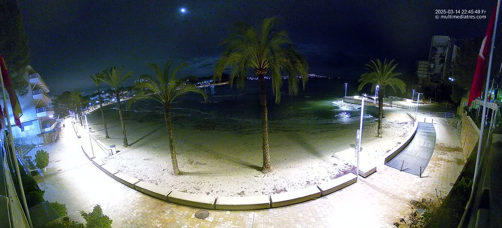 A nighttime view of a sandy beach with palm trees, a paved walkway, and buildings along the shoreline under a partly cloudy sky and a visible moon.