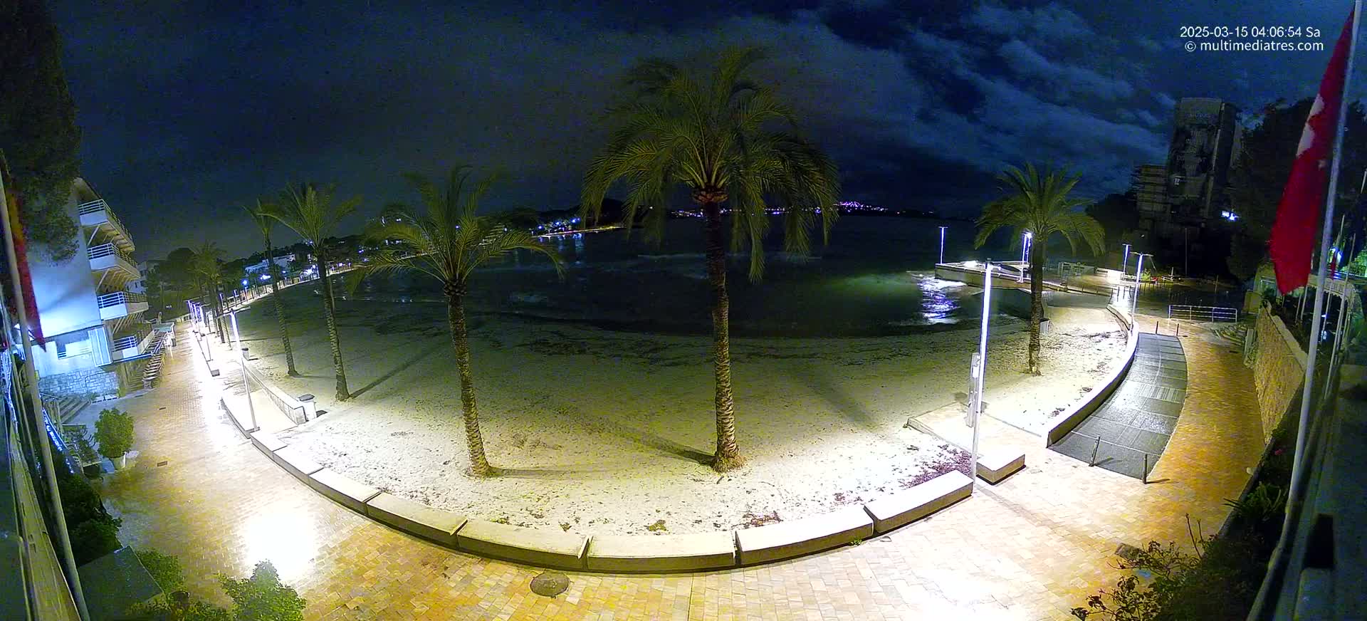 A nighttime view of a sandy beach with palm trees lining a paved walkway, illuminated by artificial lights, under a partly cloudy sky.