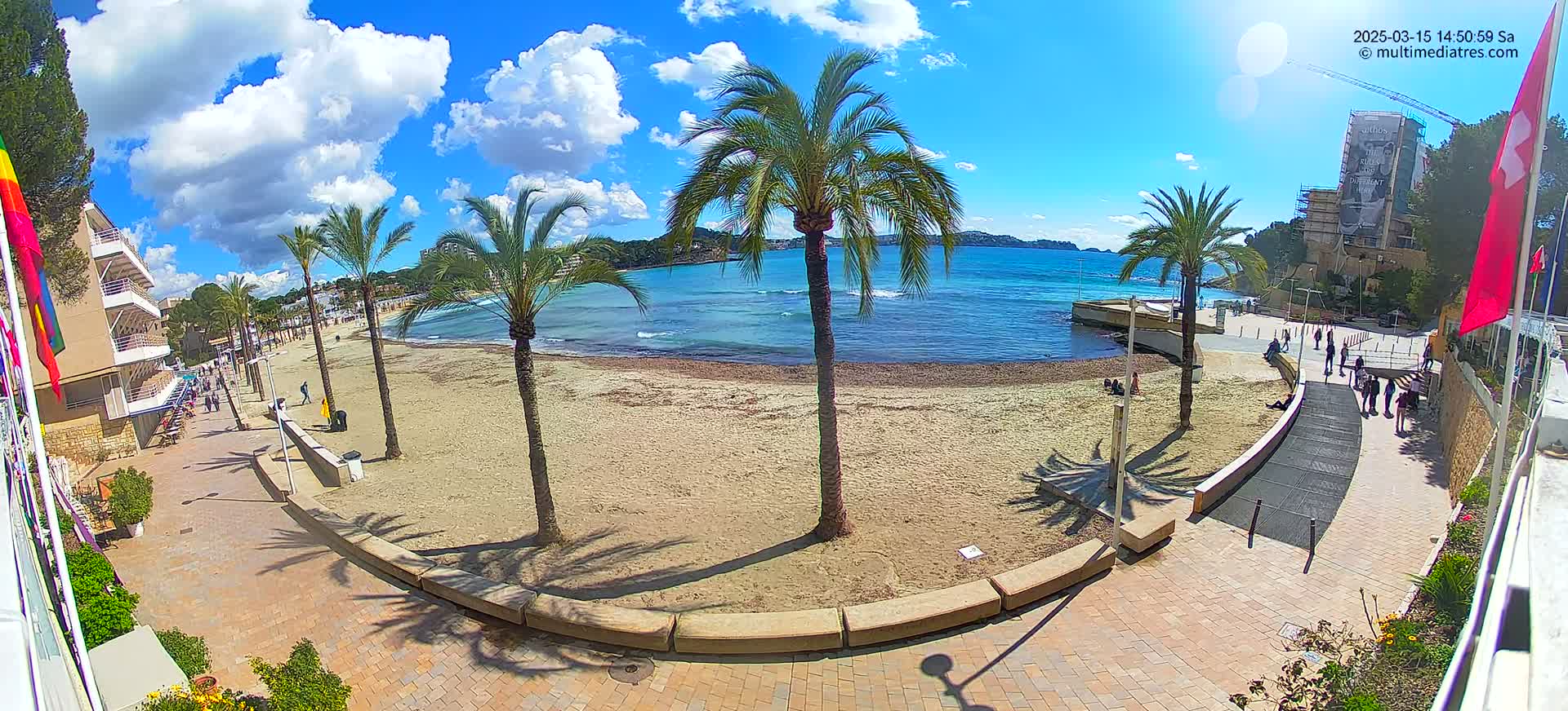 A sandy beach with palm trees and a paved walkway, overlooking a calm, blue ocean under a partly cloudy sky.