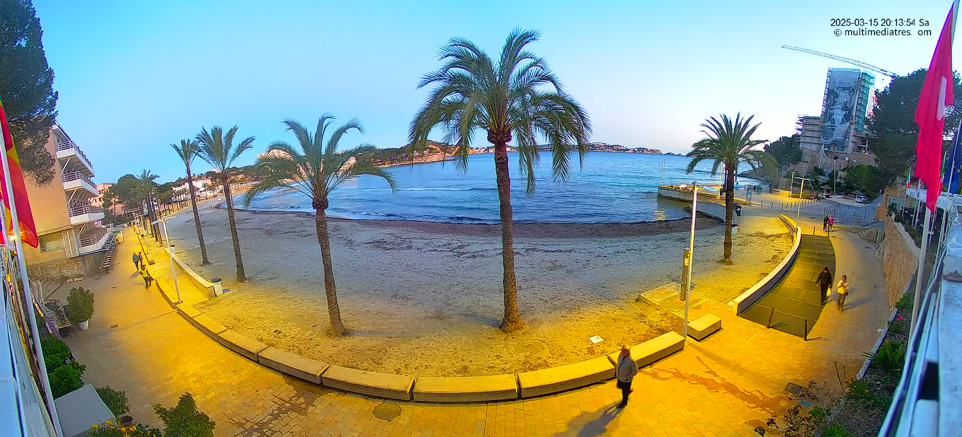 A sandy beach at twilight is bordered by a palm-lined promenade, with buildings and a calm sea in the background.