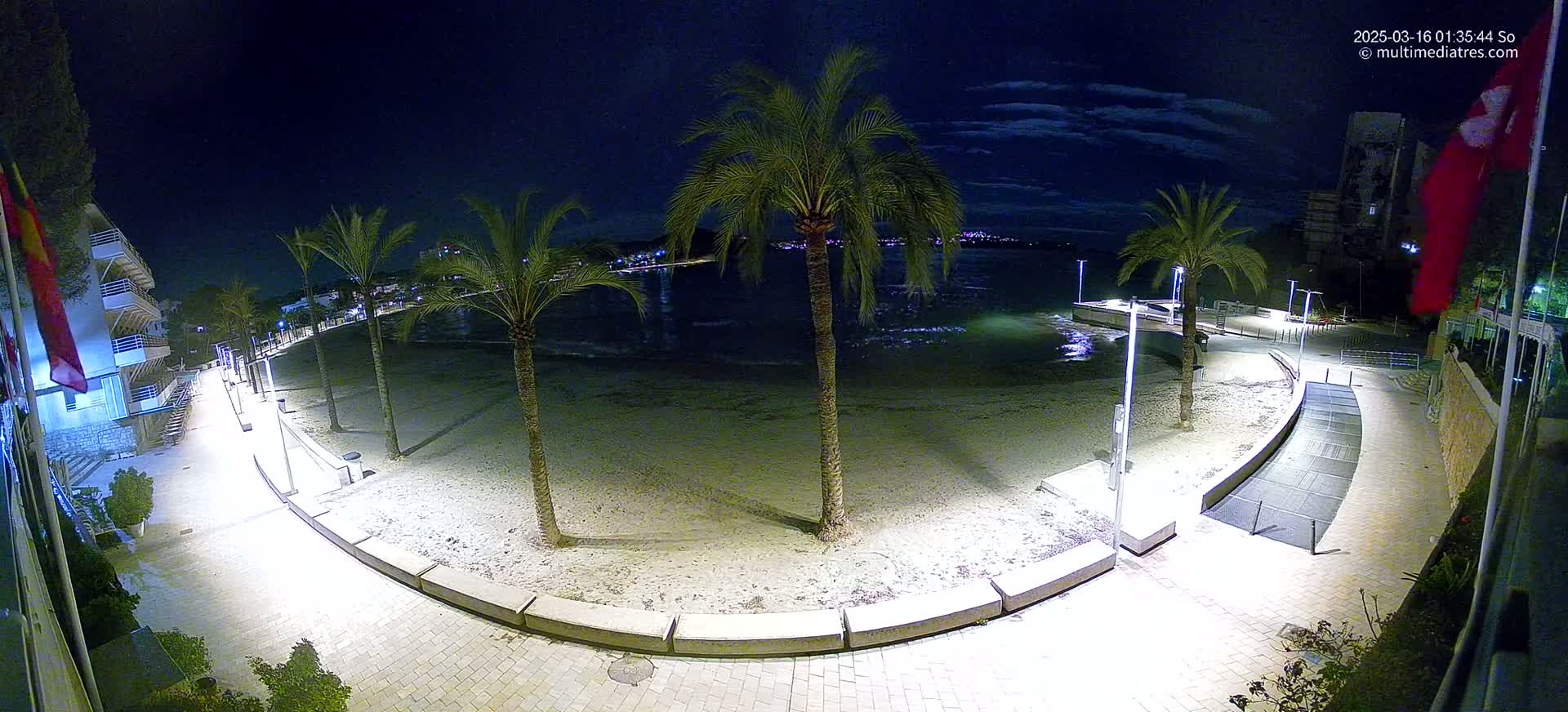 A nighttime view of a sandy beach, palm trees, and a paved walkway illuminated by lights, overlooking calm water under a partly cloudy sky.