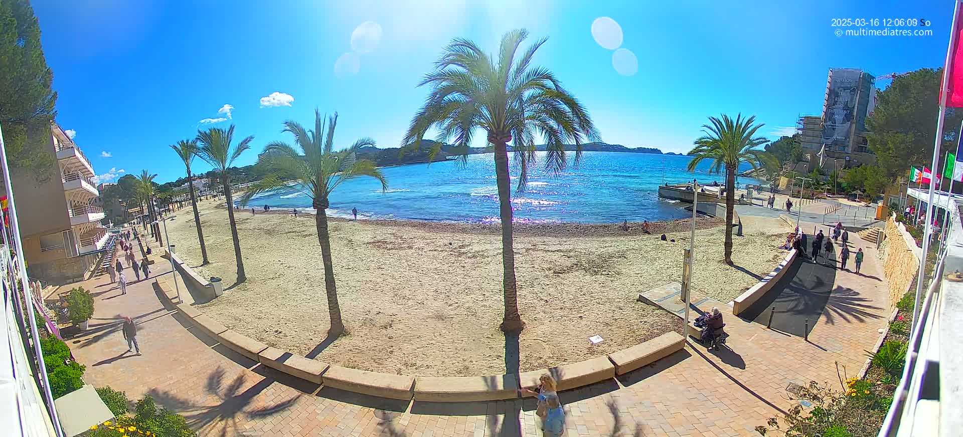 A sandy beach with palm trees lines a paved walkway next to a calm, blue ocean under a bright, sunny sky.