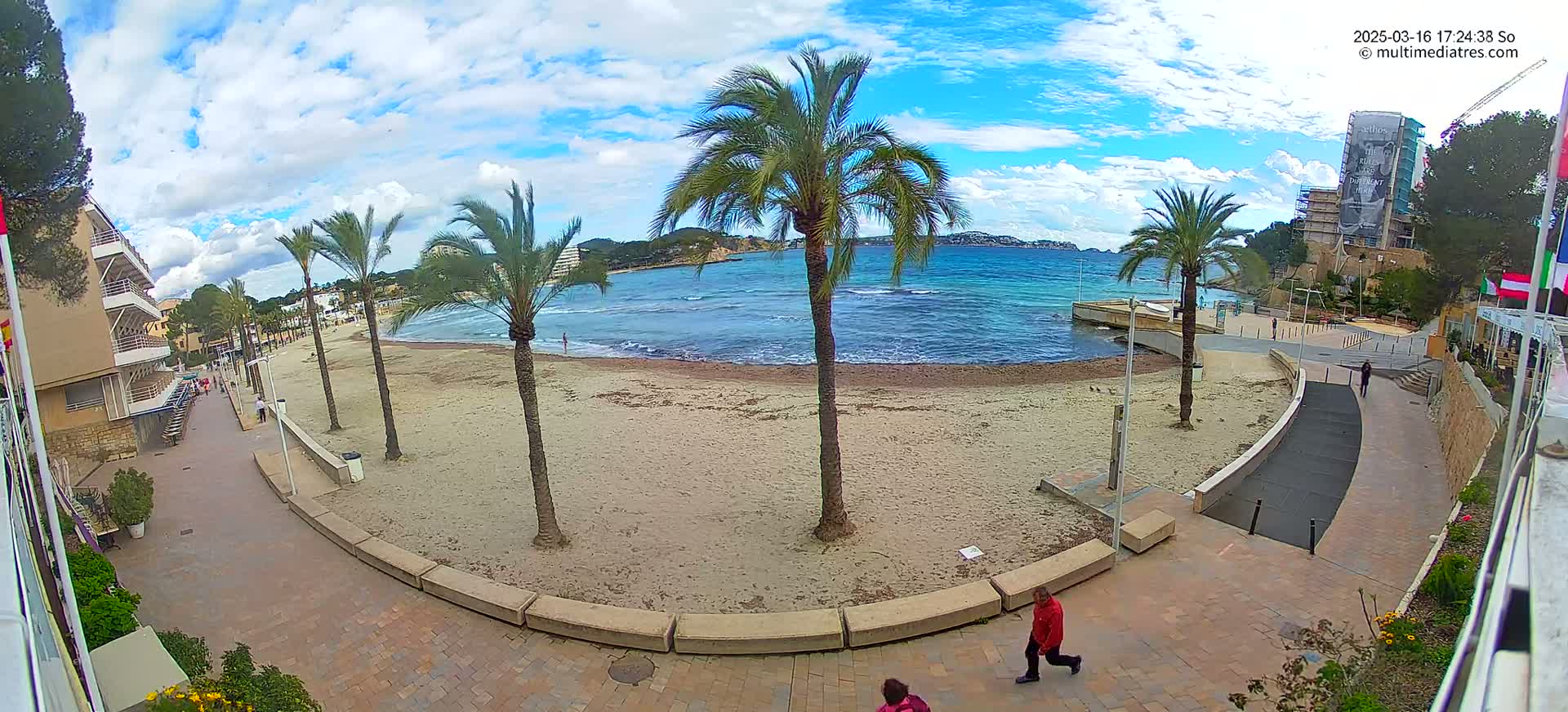 A sandy beach with palm trees and a calm, blue ocean under a partly cloudy sky is viewed from a seaside promenade.
