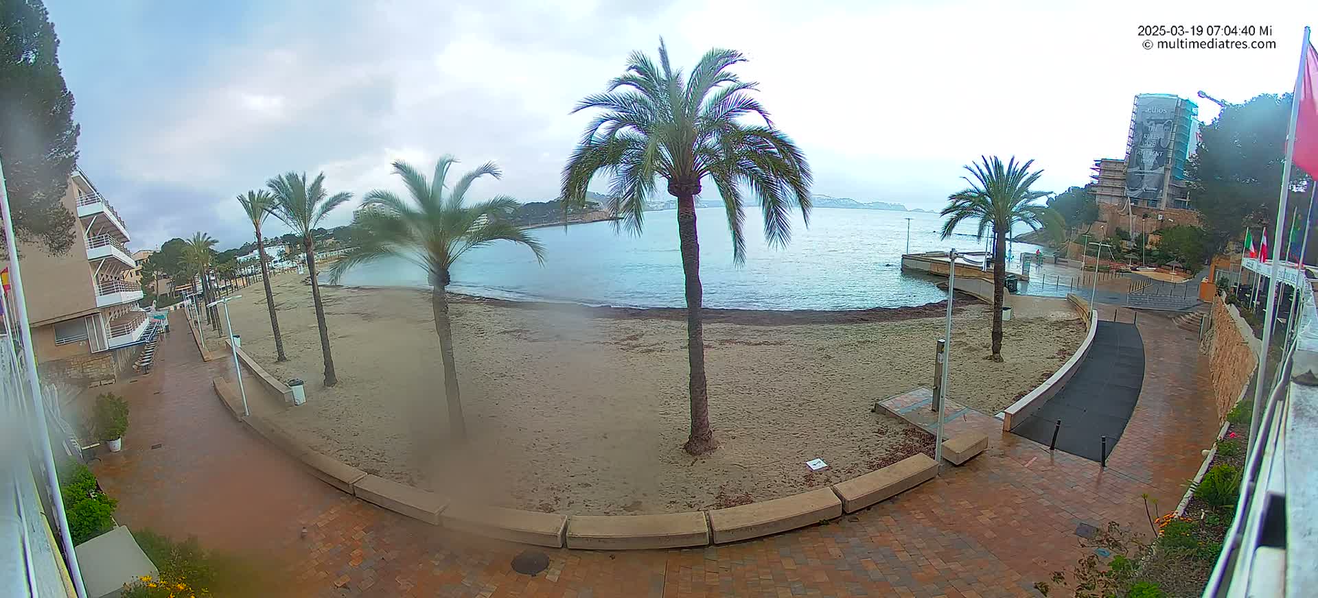 A mostly empty sandy beach with palm trees is seen on an overcast day next to a paved walkway and buildings.