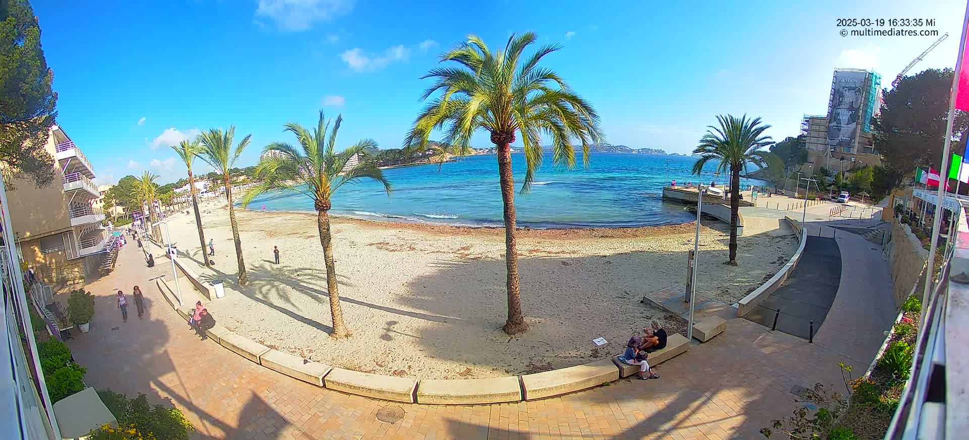 A sandy beach with palm trees and a few people on a sunny day, overlooking calm turquoise water.