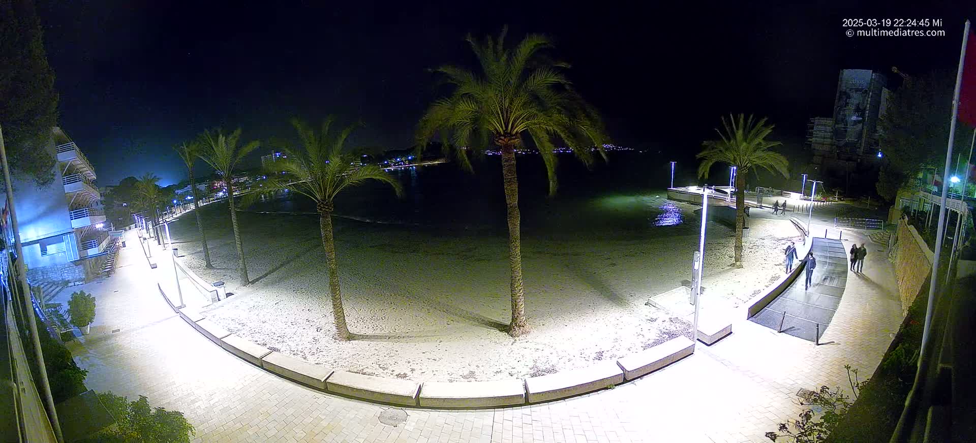 A nighttime aerial view shows a sandy beach, palm trees, a paved walkway, and a few people walking along the beachfront under clear skies.