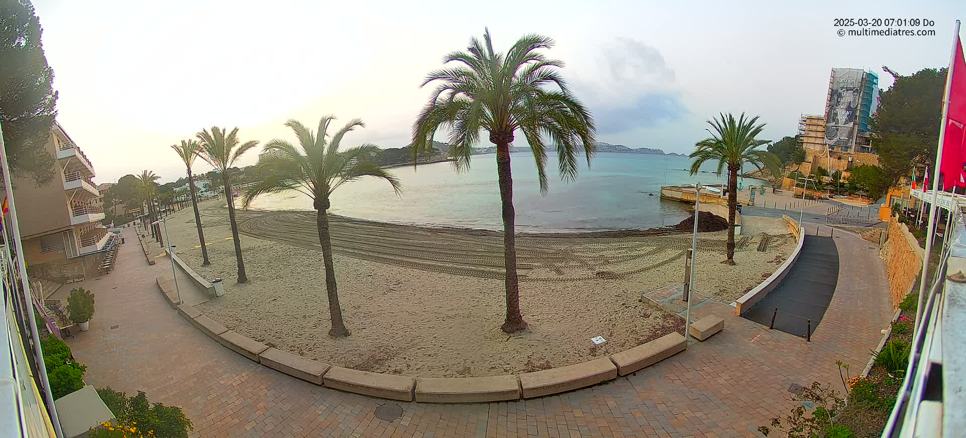 A mostly sunny day at a beach shows a sandy shoreline with tire tracks, several palm trees, and a paved walkway leading to a building under construction.