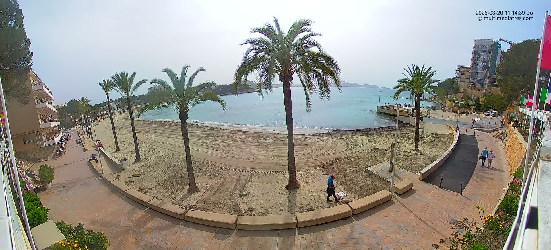 A sandy beach with palm trees and a calm sea under a bright, clear sky is viewed from above, showing a paved walkway leading to buildings and a few people walking.