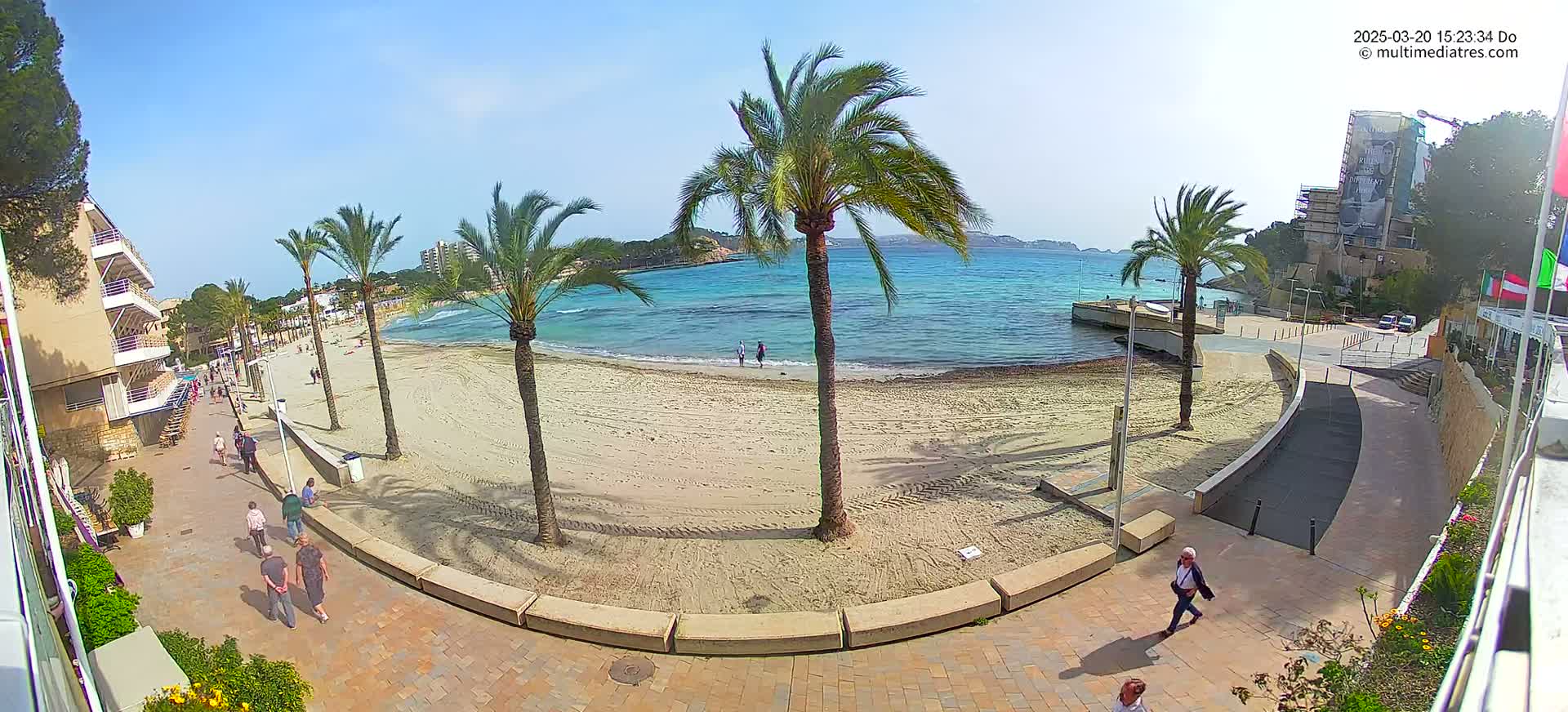 A sandy beach with palm trees and several people walking along a paved walkway next to a calm, clear blue ocean on a sunny day.