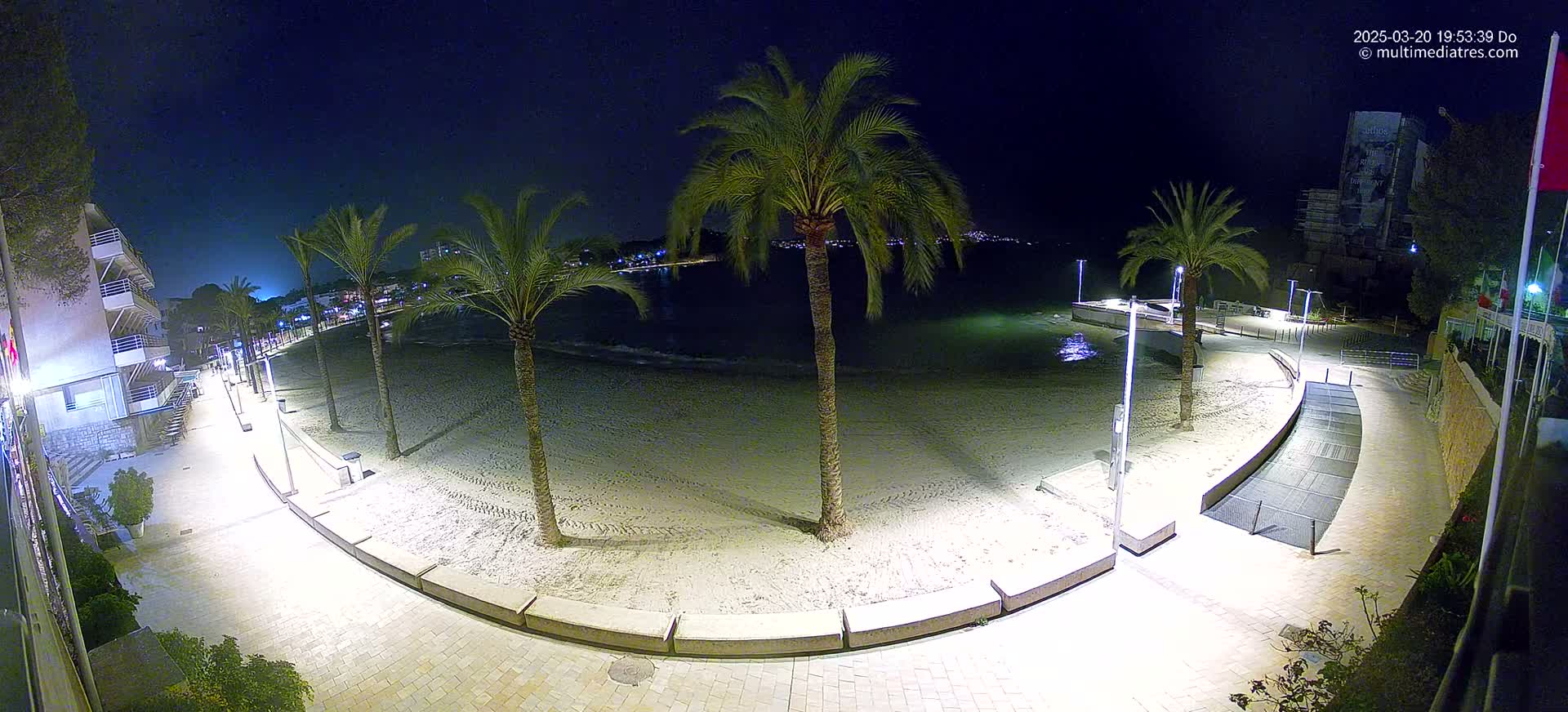 A nighttime view of a sandy beach with palm trees lining a paved walkway, illuminated by artificial lights.