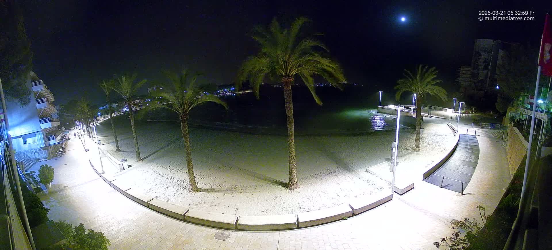 A nighttime view of a sandy beach with palm trees and a paved walkway, illuminated by lights under a mostly clear sky with a visible moon.