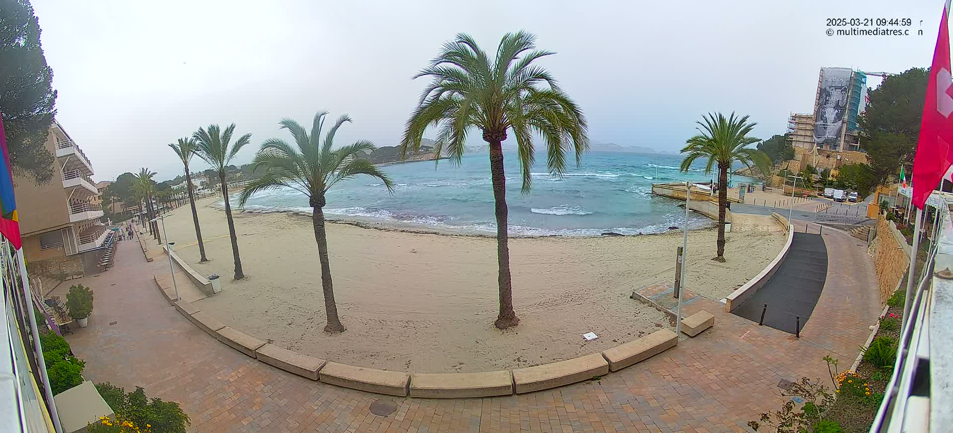 A sandy beach with palm trees and a paved walkway is visible, under an overcast sky, with a calm sea displaying small waves.