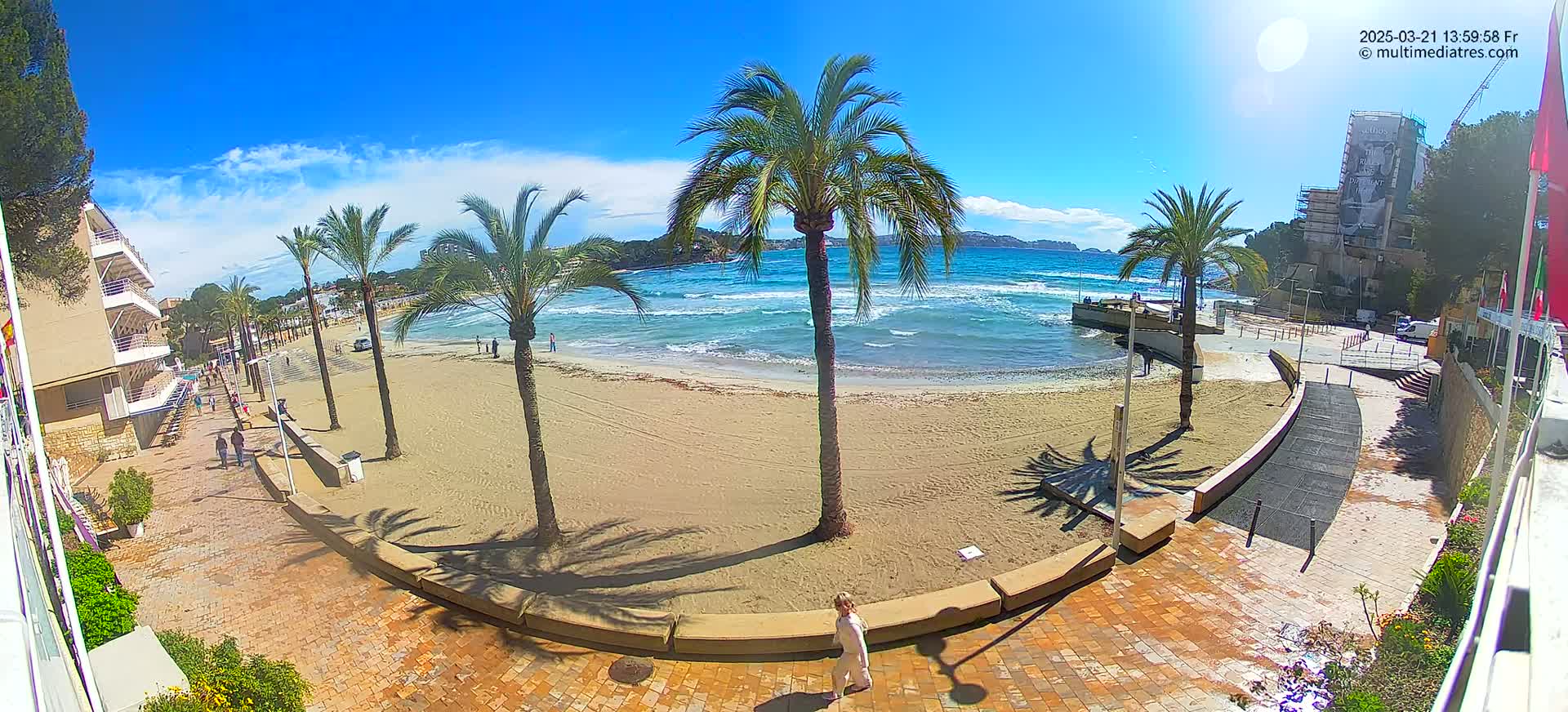 A sandy beach with palm trees, a few people, and some buildings is visible on a sunny day with some small waves in the ocean.