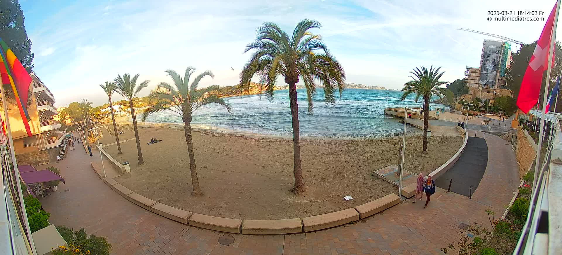A sandy beach with palm trees and a paved walkway, under a partly cloudy sky, has light waves lapping at the shore.