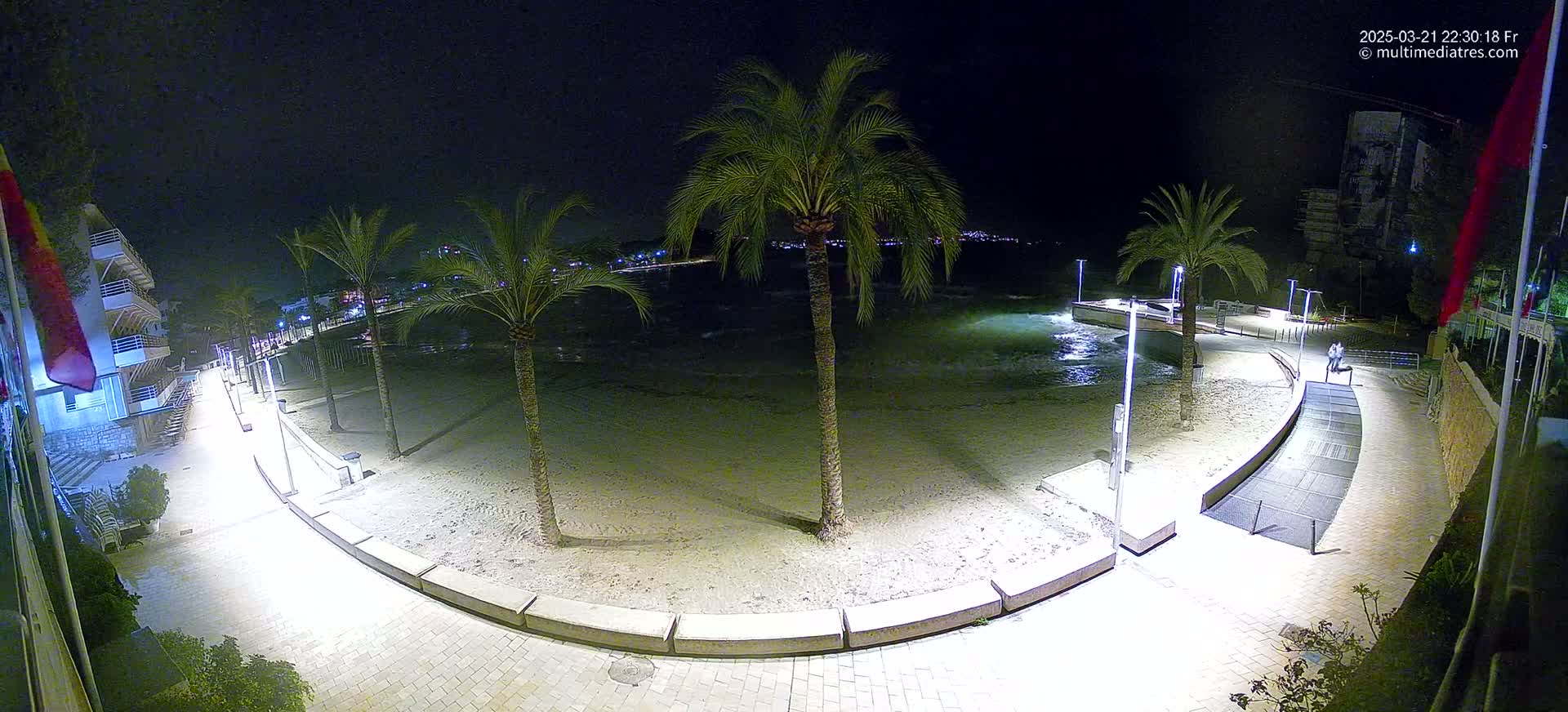 A nighttime beach scene shows a sandy shore, palm trees, and a paved walkway curving along the water's edge, illuminated by lights.