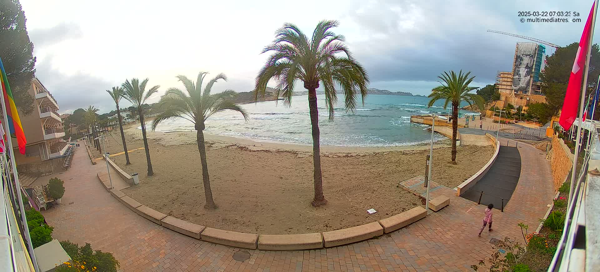A mostly cloudy day shows a sandy beach with palm trees, a paved walkway, and a person walking near a building under construction.