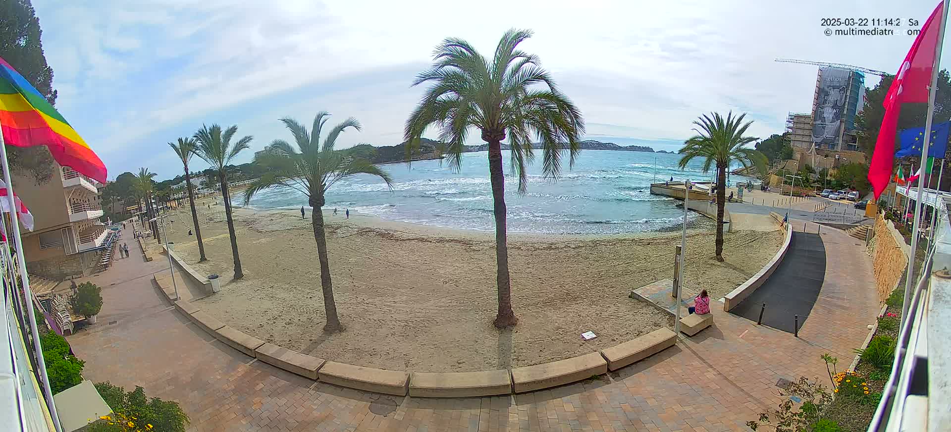 A mostly sunny day at a beach with palm trees, a sandy shore, and light waves, seen from an elevated vantage point overlooking a paved walkway and buildings.