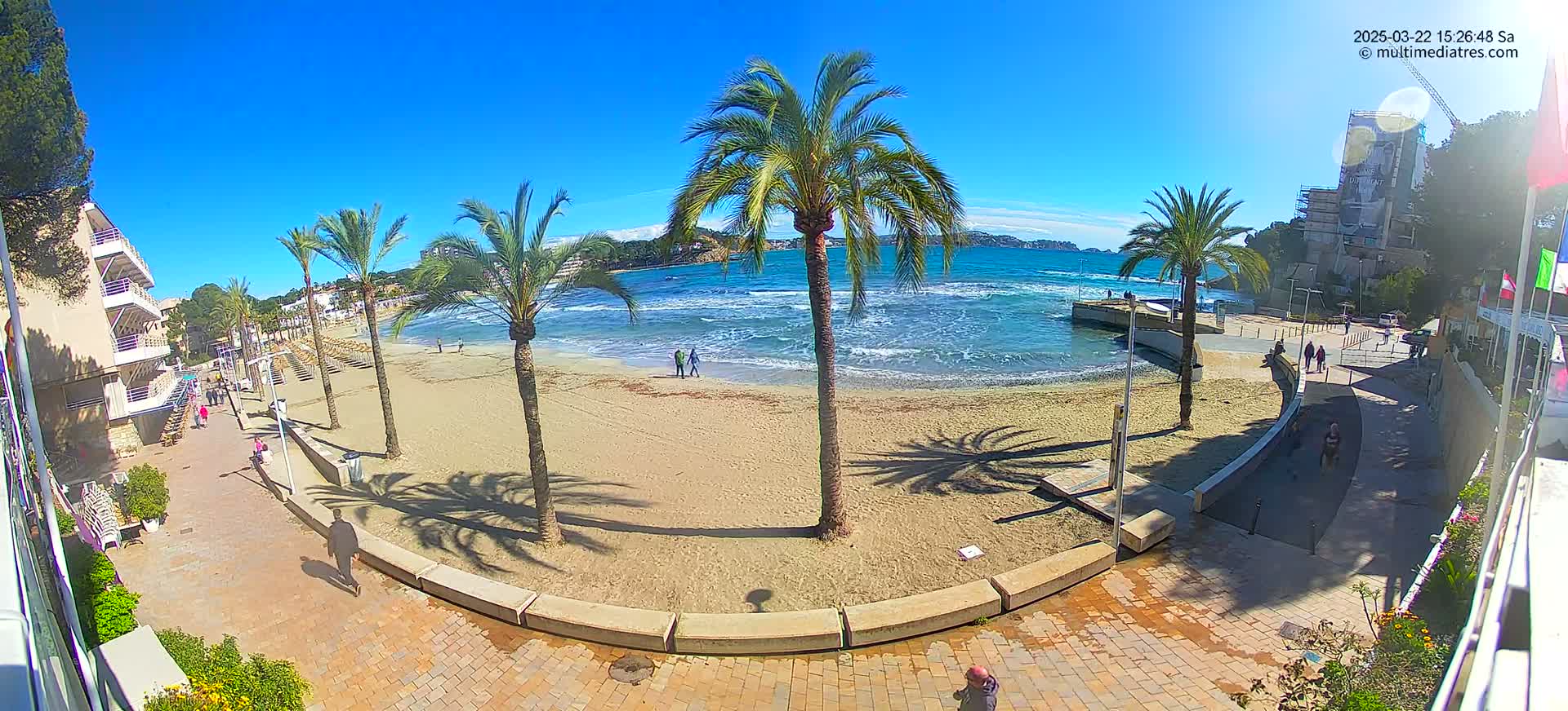 A sandy beach with palm trees and a calm, blue ocean under a bright, sunny sky is seen from a high vantage point, overlooking a paved walkway.