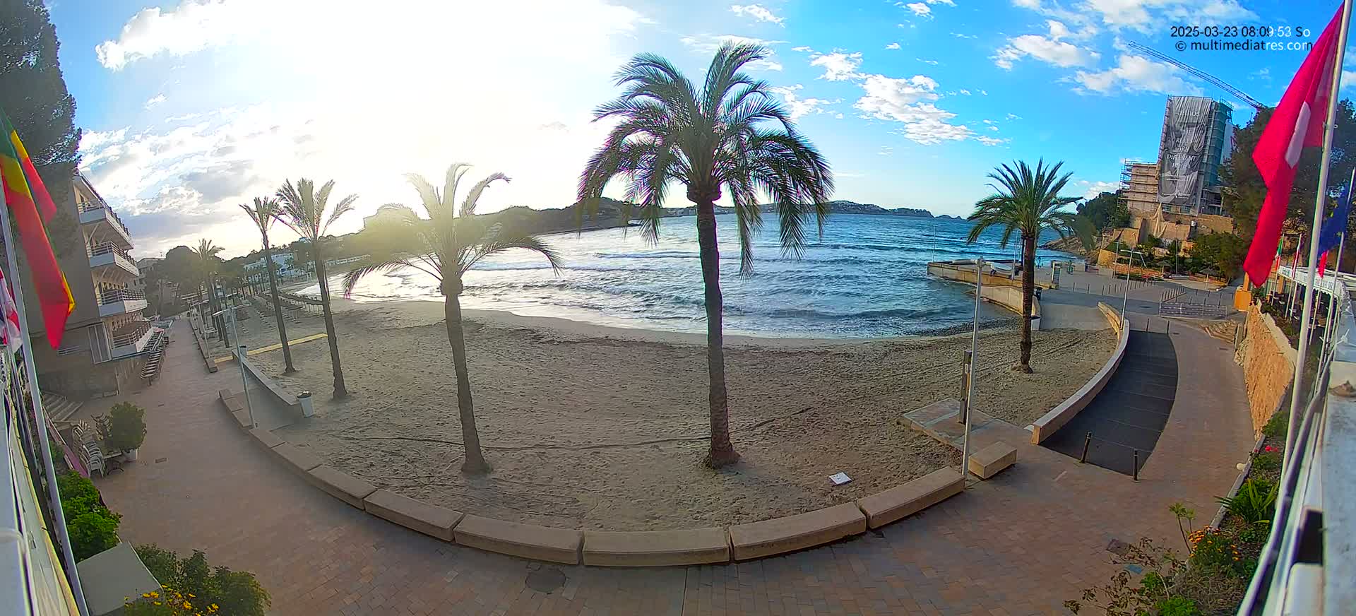 A sandy beach with palm trees is visible from a paved walkway next to a building, under a partly sunny sky with some clouds.