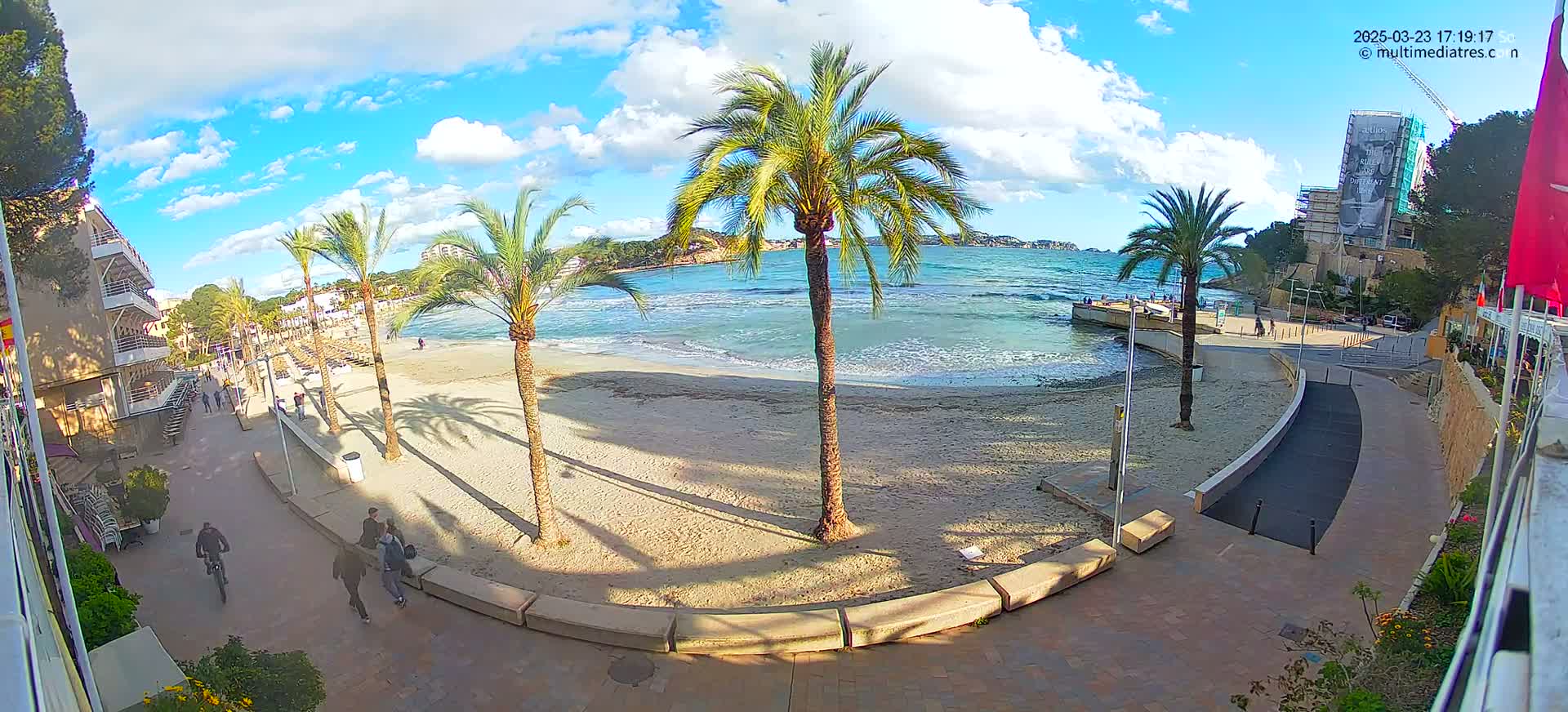A sandy beach with palm trees and a few people is next to a paved walkway, under a partly cloudy blue sky.